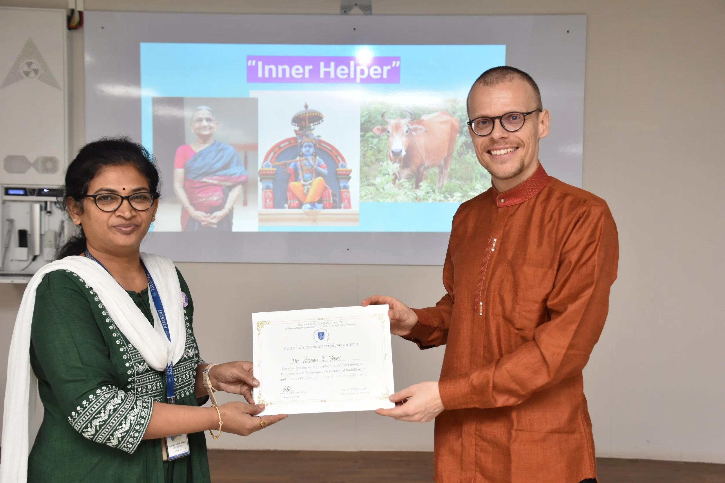 A woman in a green traditional dress and glasses receiving a certificate from a man in an orange kurta, with a presentation slide in the background titled 'Inner Helper' showing images of a woman, a Hindu deity, and a cow.