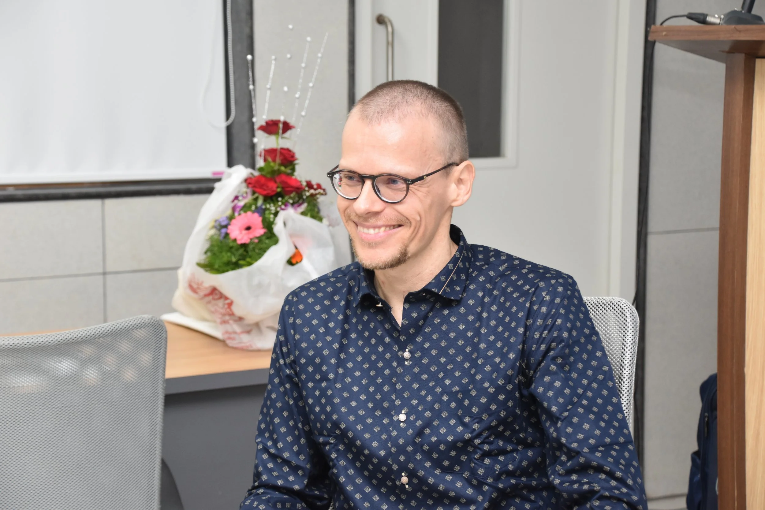 A smiling man with glasses and a trimmed beard, wearing a dark blue shirt with a geometric pattern, sitting in a modern office setting with a bouquet of flowers on the desk behind him.