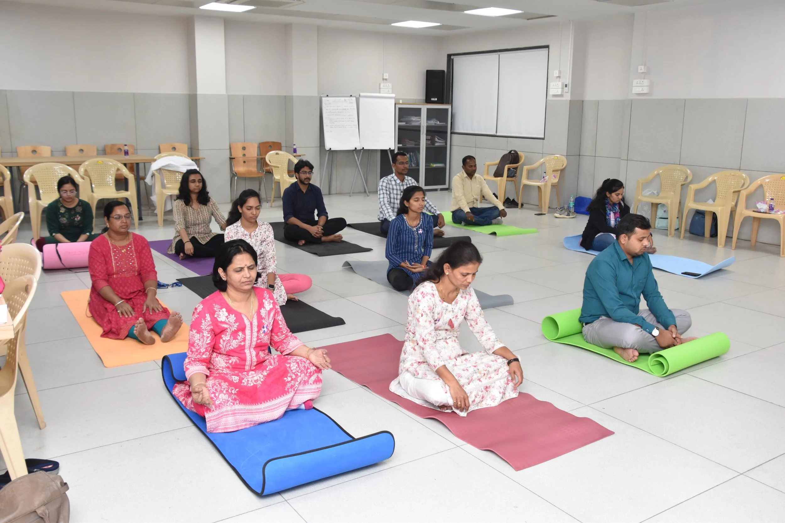 People participating in a group yoga or meditation class in a spacious room, sitting cross-legged on yoga mats with eyes closed, in a meditative pose.
