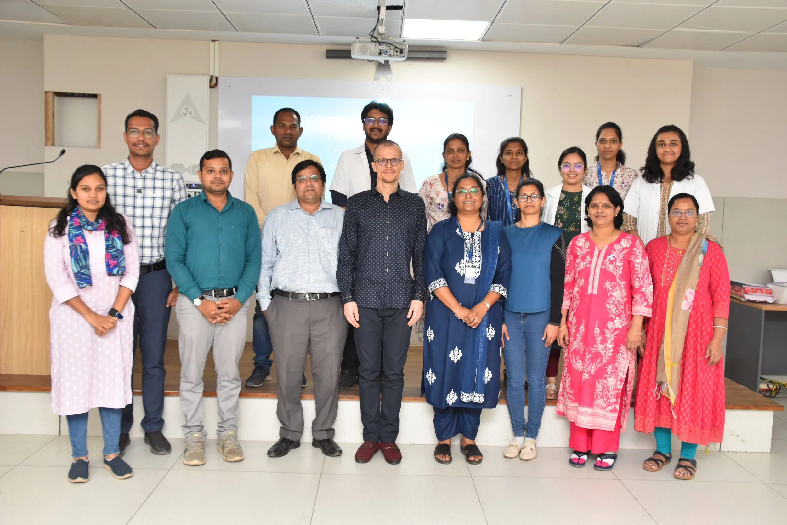 A group of 16 diverse professionals posing together in a classroom or conference room with a projector and screen behind them.