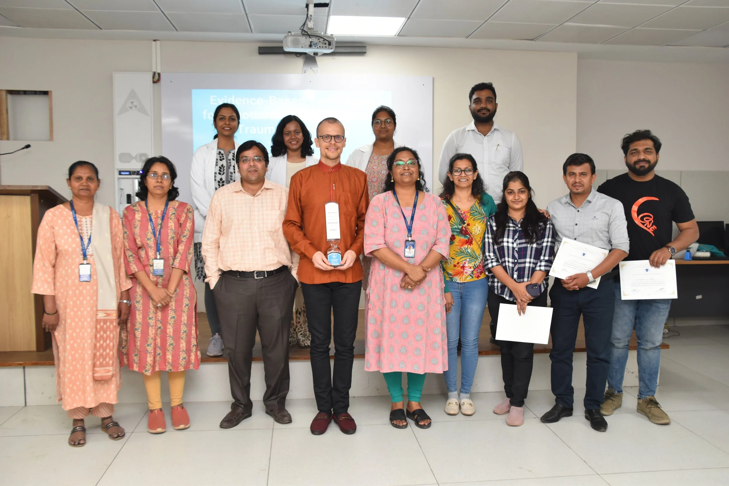 Group of people in a classroom, some holding certificates, posing for a photo after a presentation or award ceremony.