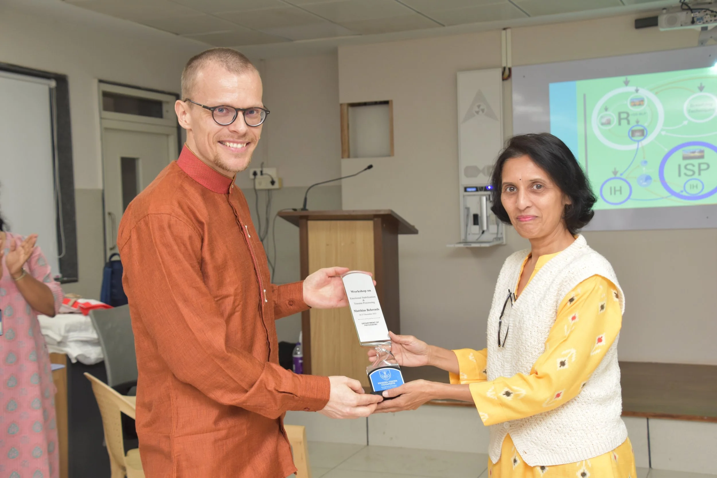 A man in an orange traditional outfit receives a plaque and a medal from a woman in a yellow dress and white vest during an indoor event, with a presentation slide in the background.