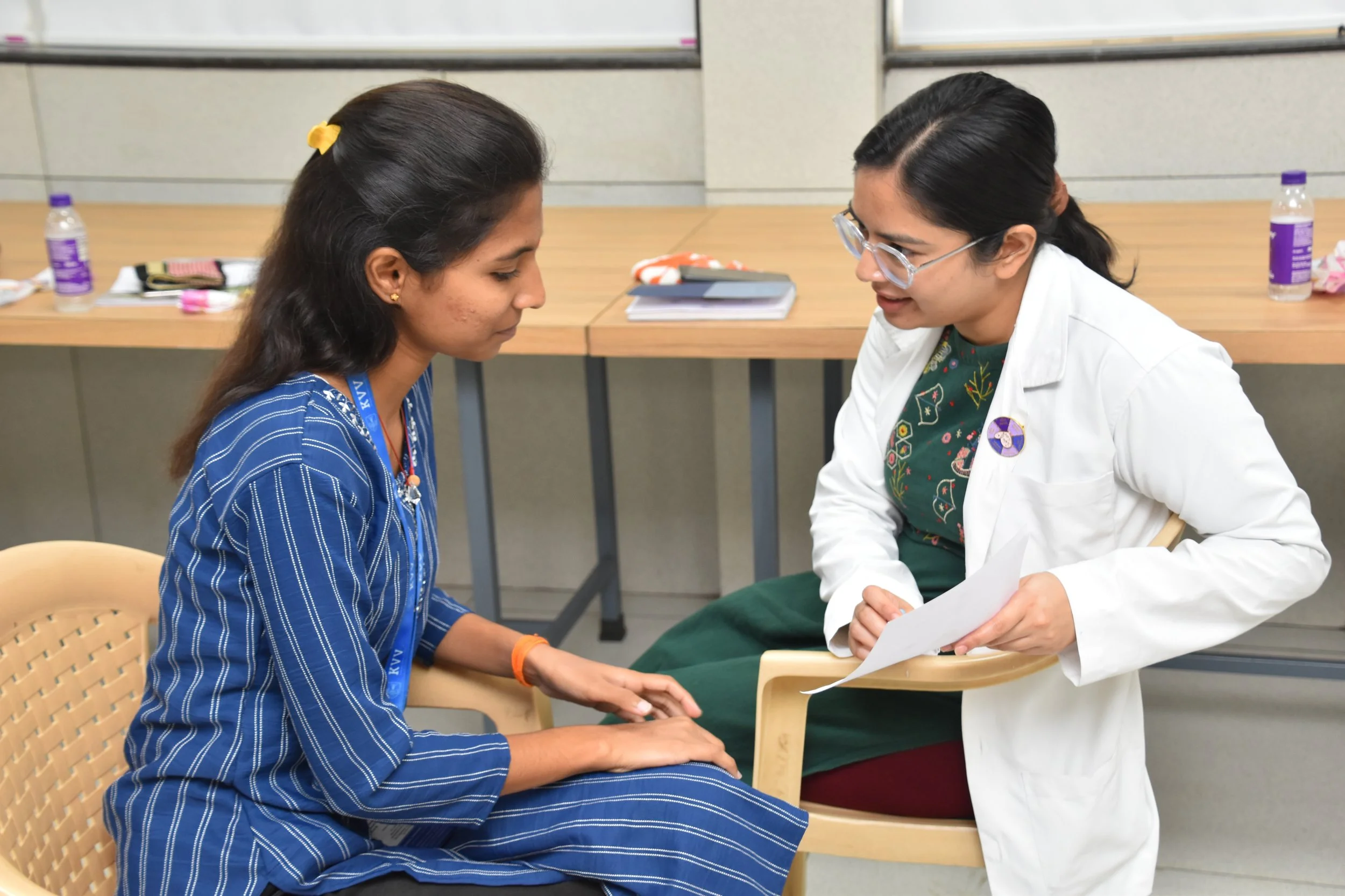 Two women having a conversation in a clinical setting, one sitting with hands on her lap and the other leaning forward holding a piece of paper, both engaged in a discussion.