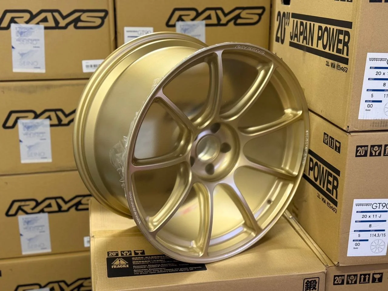 Golden alloy wheel rim placed on a cardboard box in front of stacked boxes labeled RAYS and JAPAN POWER.