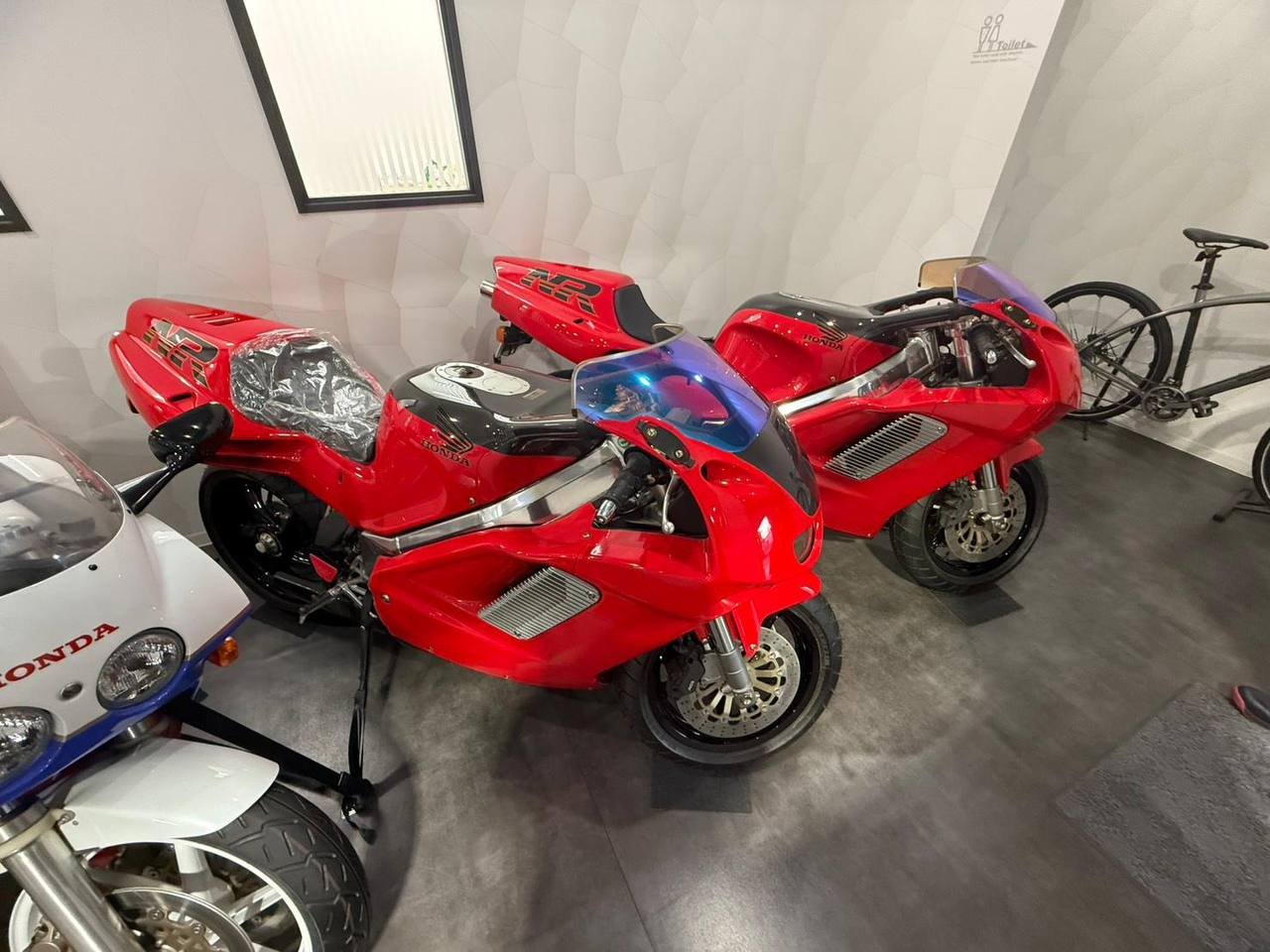 Two red Honda sport motorcycles parked indoors, with a white and blue Honda motorcycle and two bicycles visible nearby.