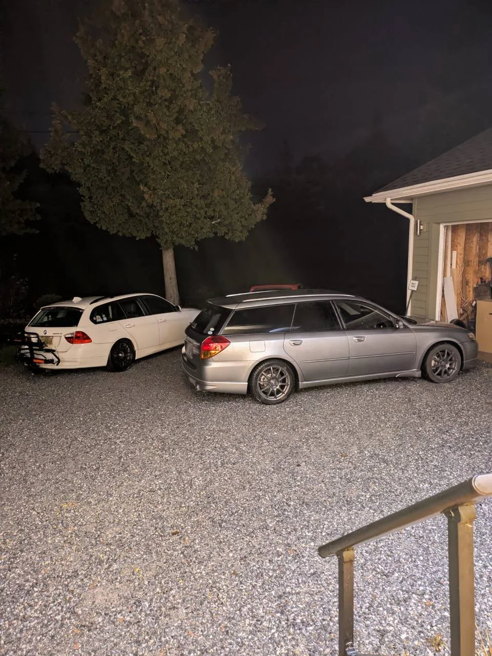 Two cars parked in a gravel driveway next to a house with an open garage. There is a large tree and a dark sky in the background at night.