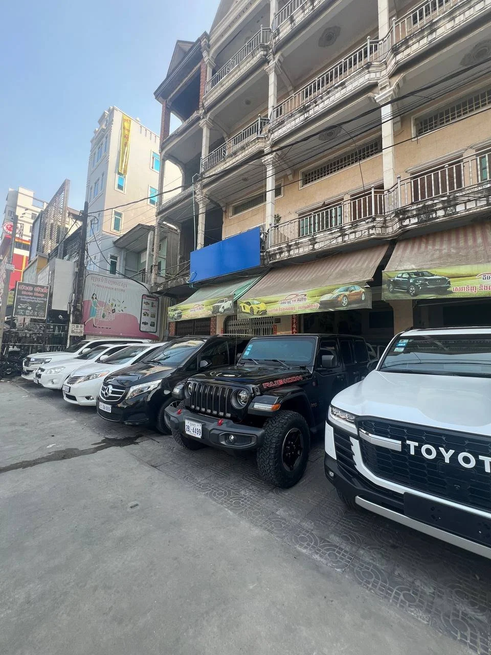 Street view showing parked cars in front of multi-story buildings with balconies, including a black Jeep Rubicon and a white Toyota. Buildings have signs and awnings, some with advertisements for cars and others with a blank blue sign.