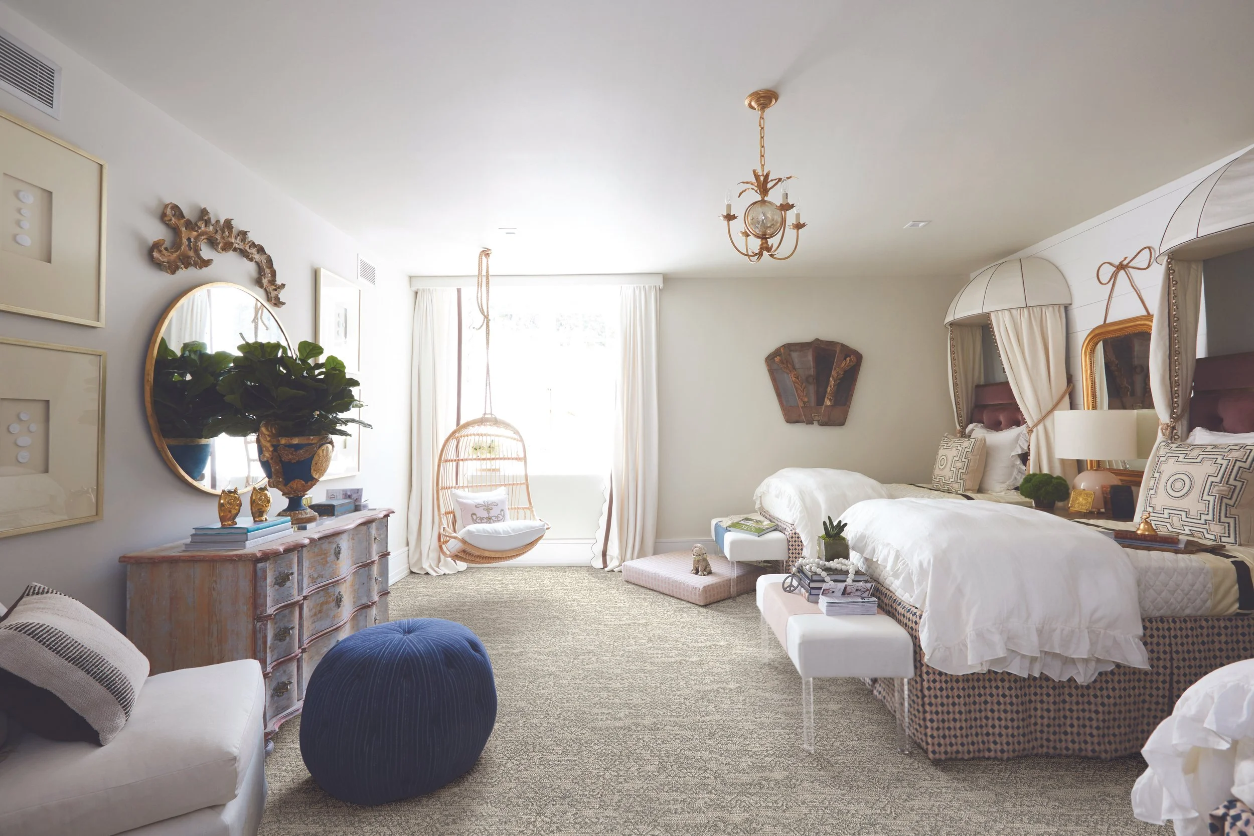 A bright, elegantly decorated bedroom with white walls, a white ceiling, and a beige carpet. There are two beds with canopy frames, multiple pillows, and a nightstand with decorative items. A large window with white curtains lets in sunlight. The room features gold accents, a vintage chandelier, a round mirror, framed artwork, a large potted plant, and various decorative objects on furniture and shelves.