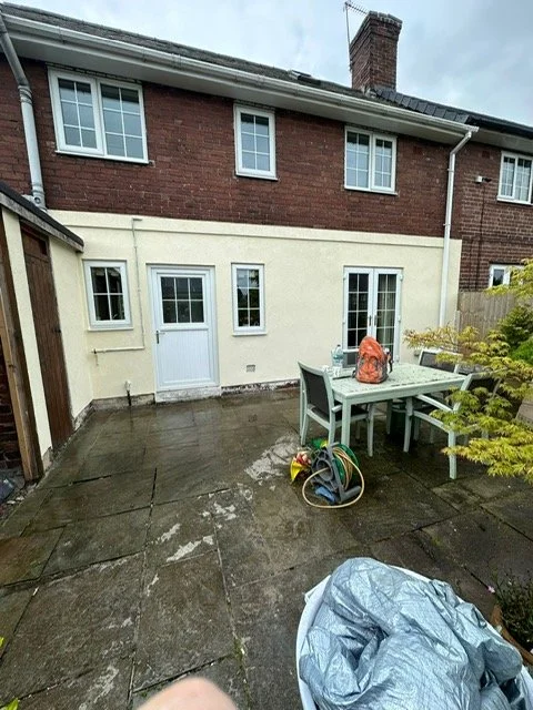 Backyard patio with a table, chairs, garden hose, and a tarp on wet concrete, with part of a two-story brick house and trees in the background.