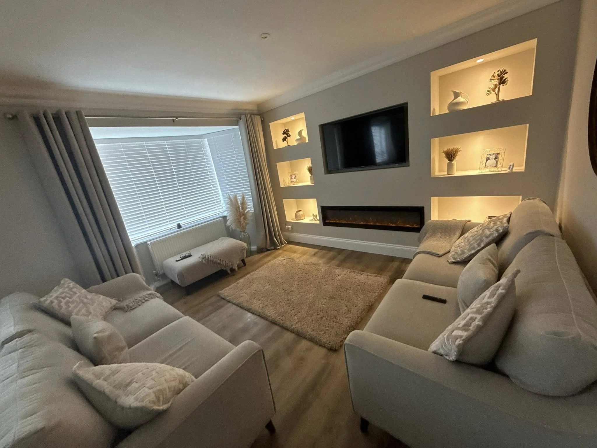 Living room with two white sofas, a rectangular area rug, a wall-mounted flat-screen TV, built-in wall shelves with decorative items, a window with blinds and curtains, and a small upholstered bench near the window.