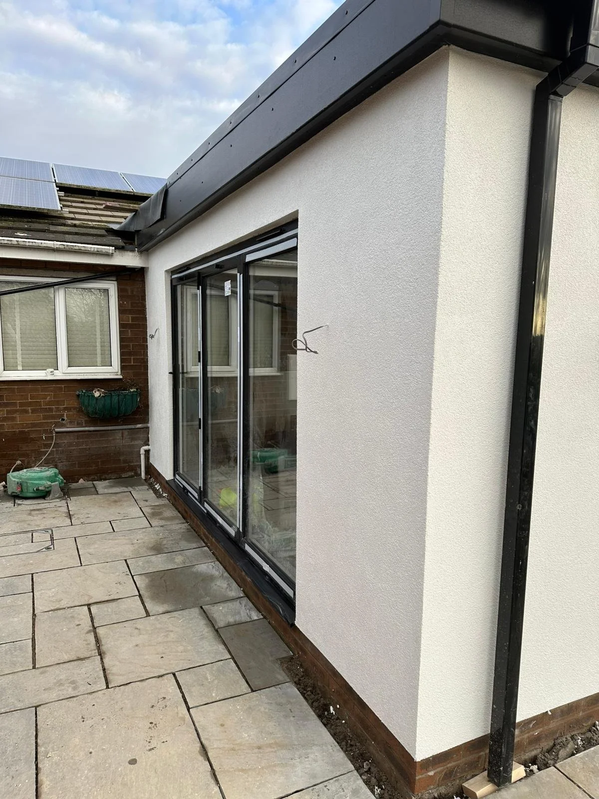 Exterior of a building with a newly installed sliding glass door, a white textured wall, and a paved patio area. Some construction materials are visible.