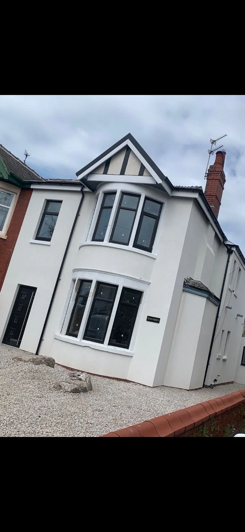 A white two-story house with black window frames and a bay window on the front. The house has a gable roof with black and white decorative trim, and a brick chimney on the right side. The front yard is covered with gravel, and there is a small brick border at the front. The sky above is cloudy.