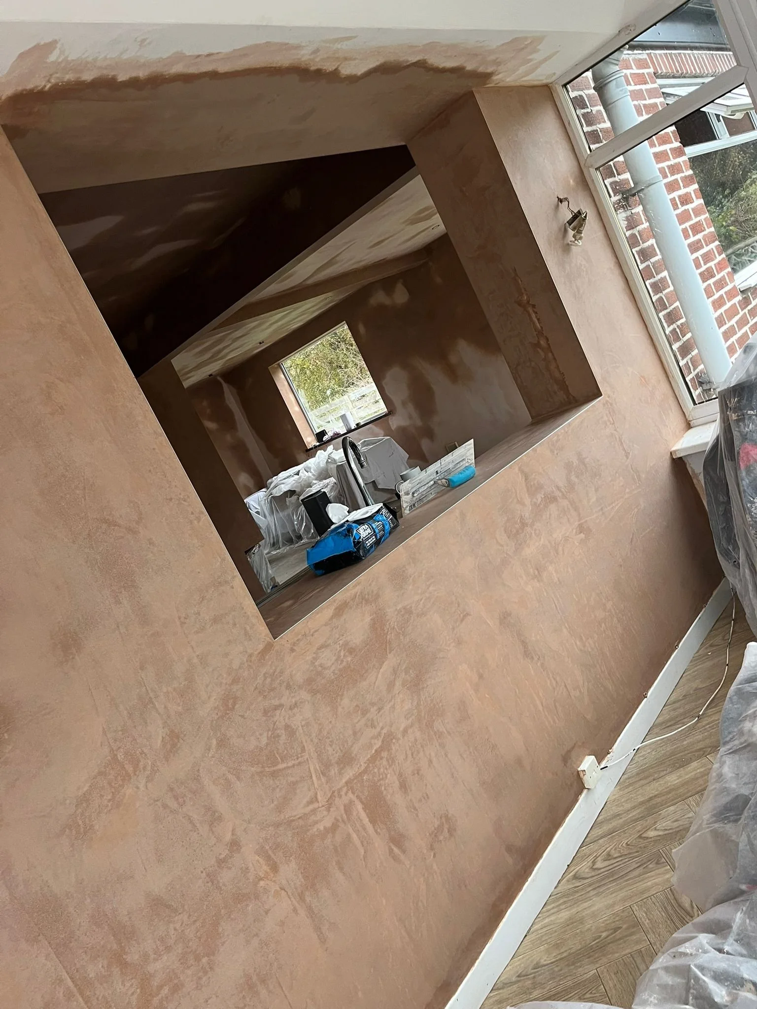 Interior of a room under renovation with walls partially painted in a warm beige color, a window revealing an outdoor view, and construction materials on a ledge.