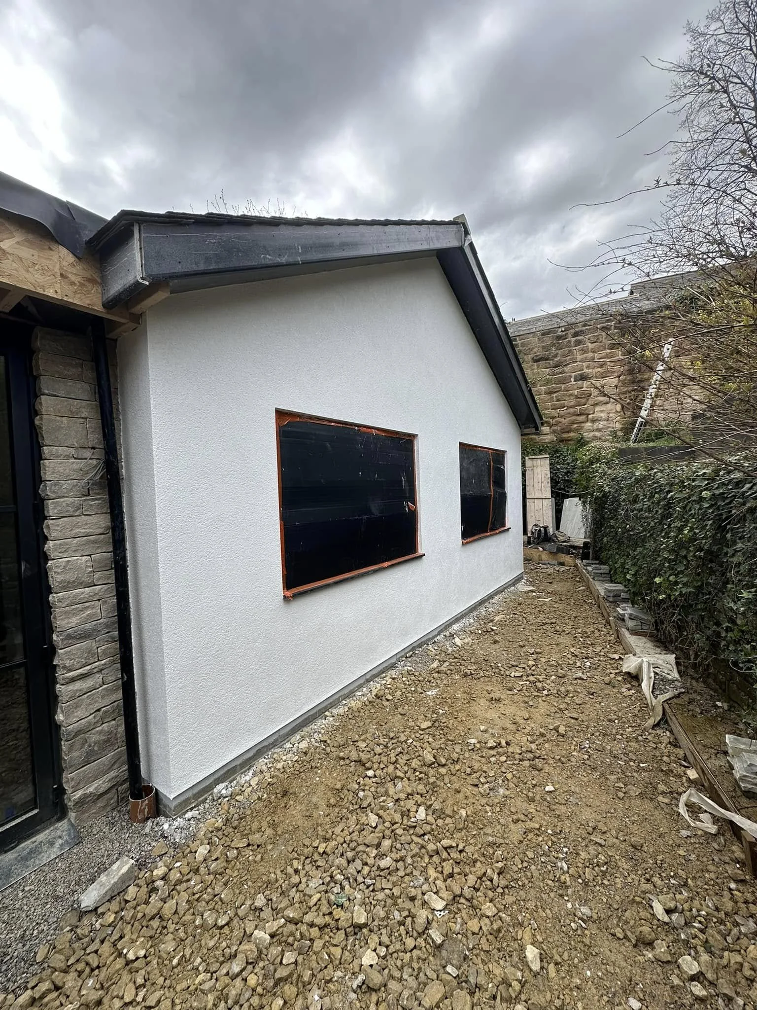 Exterior of a house under construction with a freshly rendered white wall, two covered window openings, and an unfinished ground area with rocks and dirt.