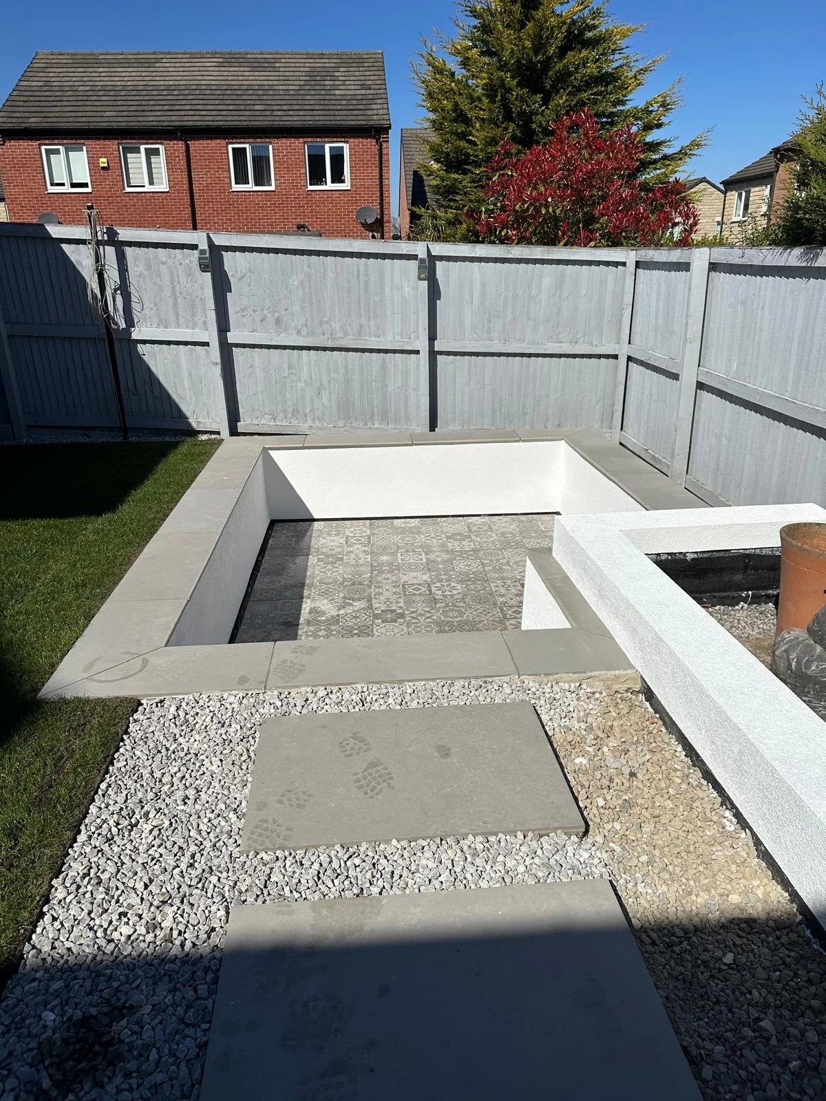 A freshly rendered sunken patio with a patterned tile bottom, surrounded by a white render border, fences, and a small patch of grass on the left. The sky is clear and blue.
