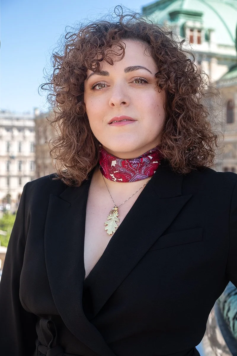 Eine Frau mit curly braunen Haaren, schwarzer Jacke, roter Halstuch, und einer goldenen Baum-Halskette auf einem Stadtbild im Hintergrund an einem sonnigen Tag in Wien vor der Staatsoper