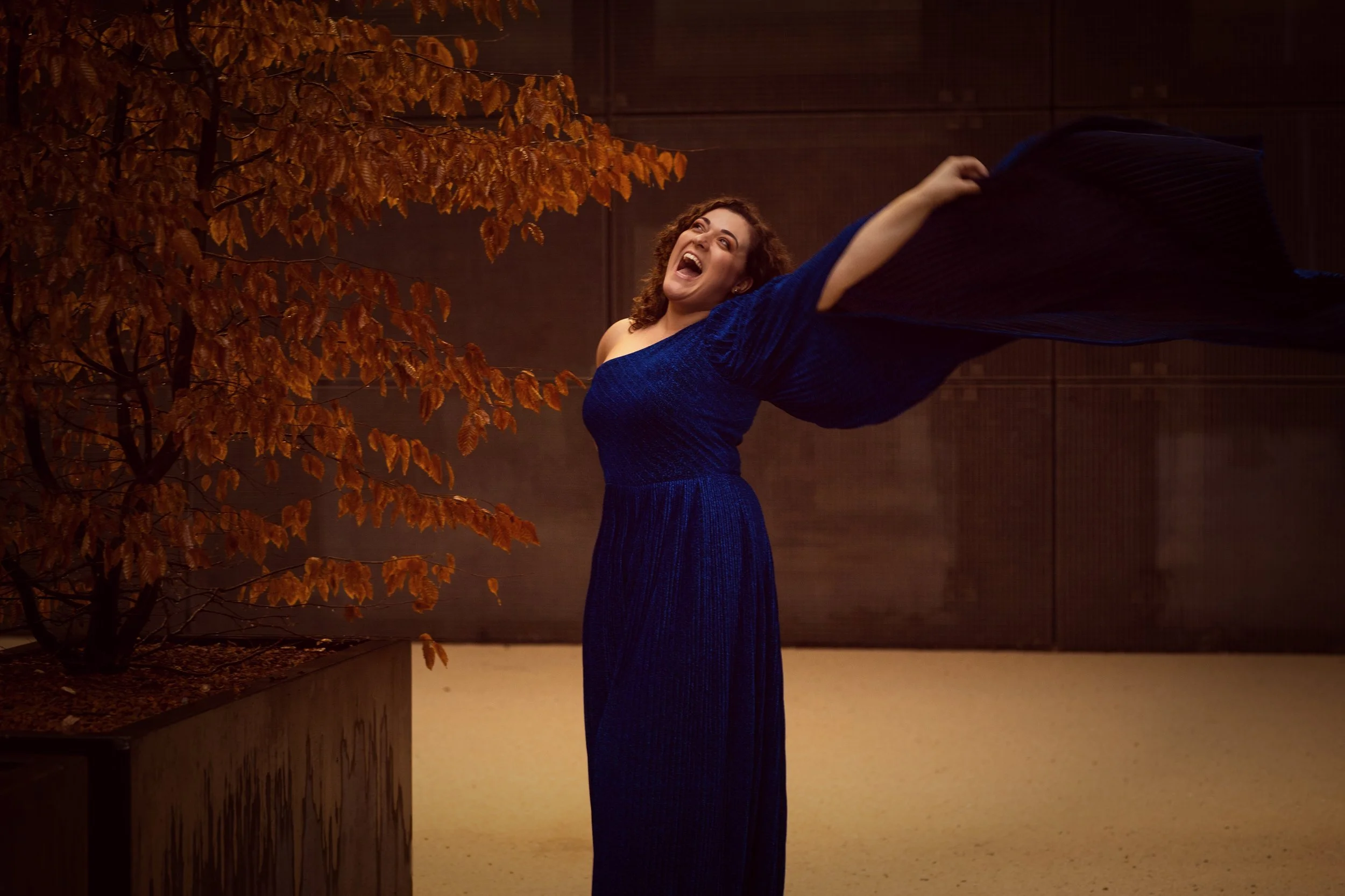 Frau in einem blauen Kleid, die vor einem Baum mit orangefarbenen Herbstblättern steht und laut singend die Arme ausbreitet.