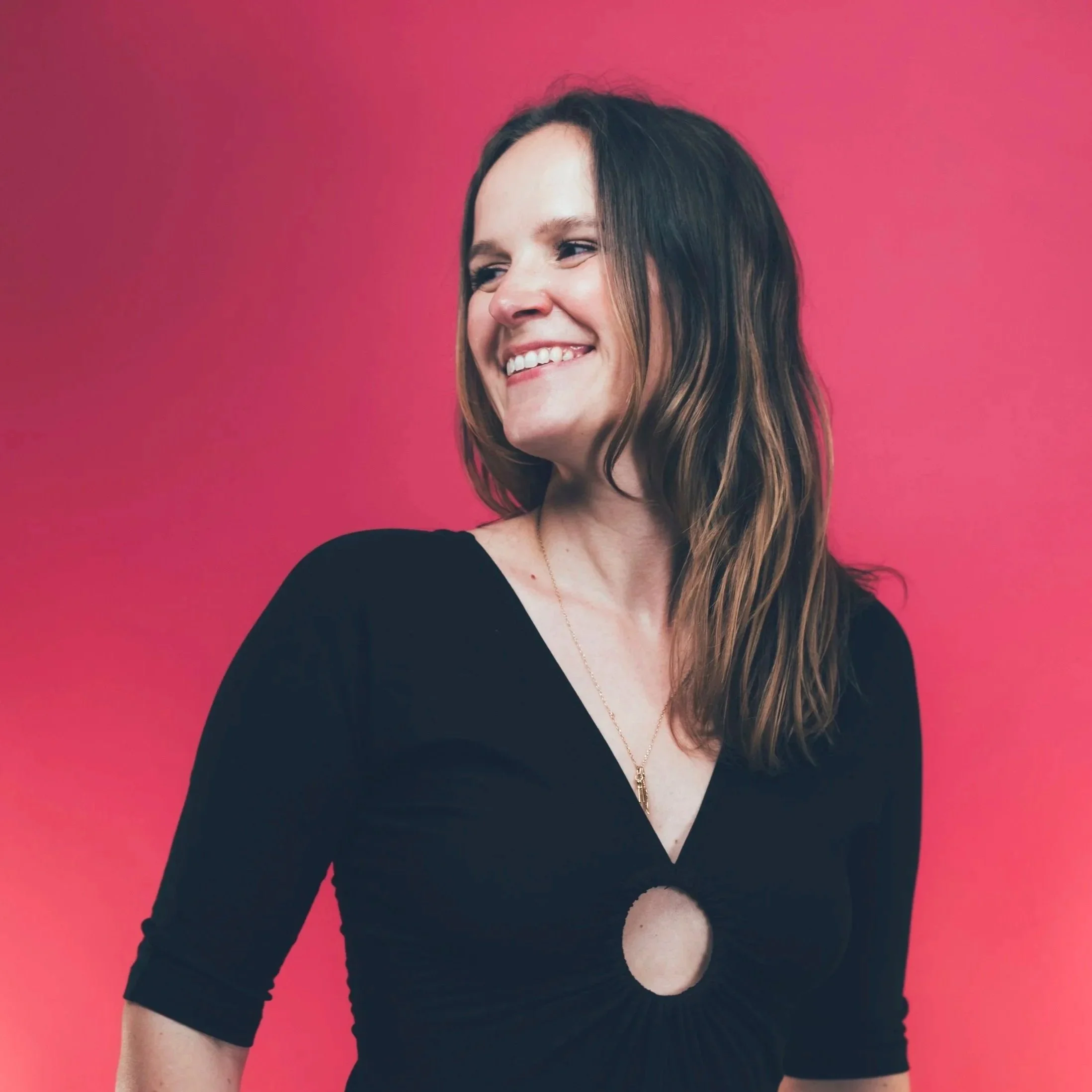 A woman with shoulder-length brown hair smiling and looking to her left, wearing a black top with a circular cutout in the front and a gold necklace, against a pink background.