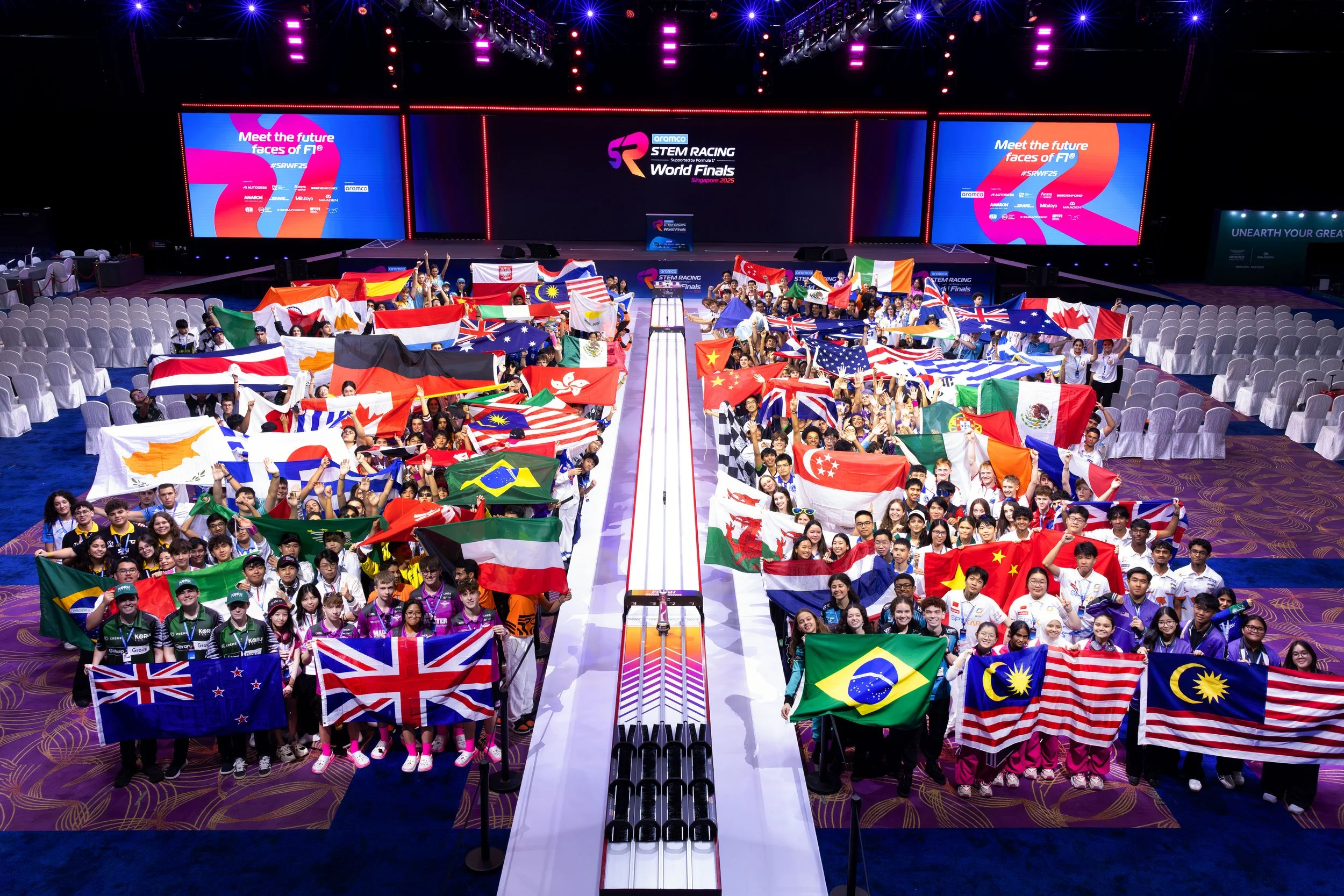 Groups of young athletes holding various national flags inside a large indoor stadium with a racing track in the center, stage screens displaying event information, and spectators in the background.