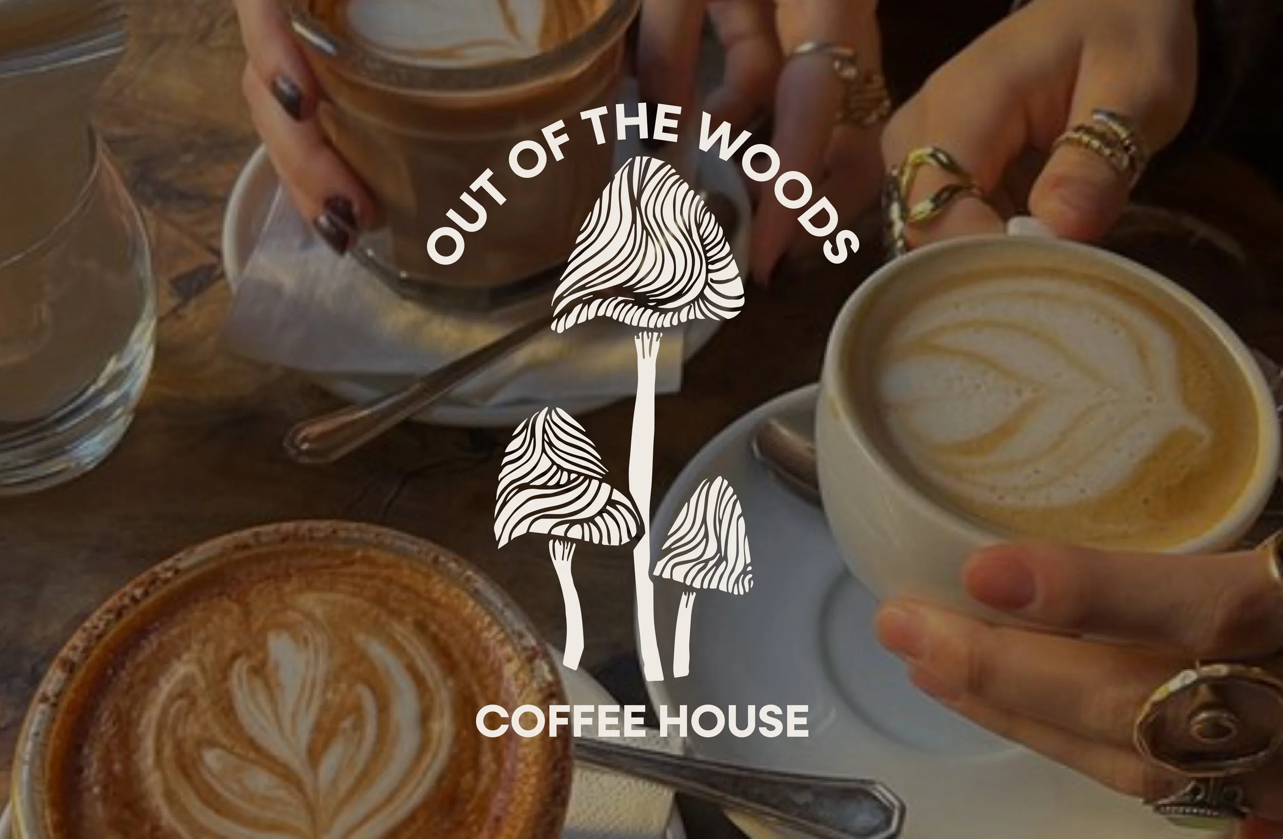 People holding cups of coffee with latte art, on a wooden table at a coffee shop. The shop's logo, Out of the Woods Coffee House, is overlaid on the image.