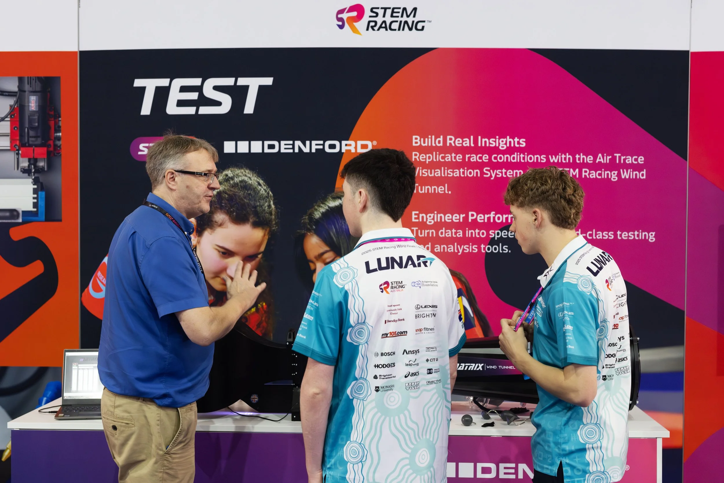 Two young male students in blue racing shirts engaged in conversation with an older man in a blue polo shirt at a STEM Racing exhibition booth. Behind them, a large display screen shows promotional content for STEM Racing, with text describing build insights and testing processes. The booth features various technology and racing-related materials.