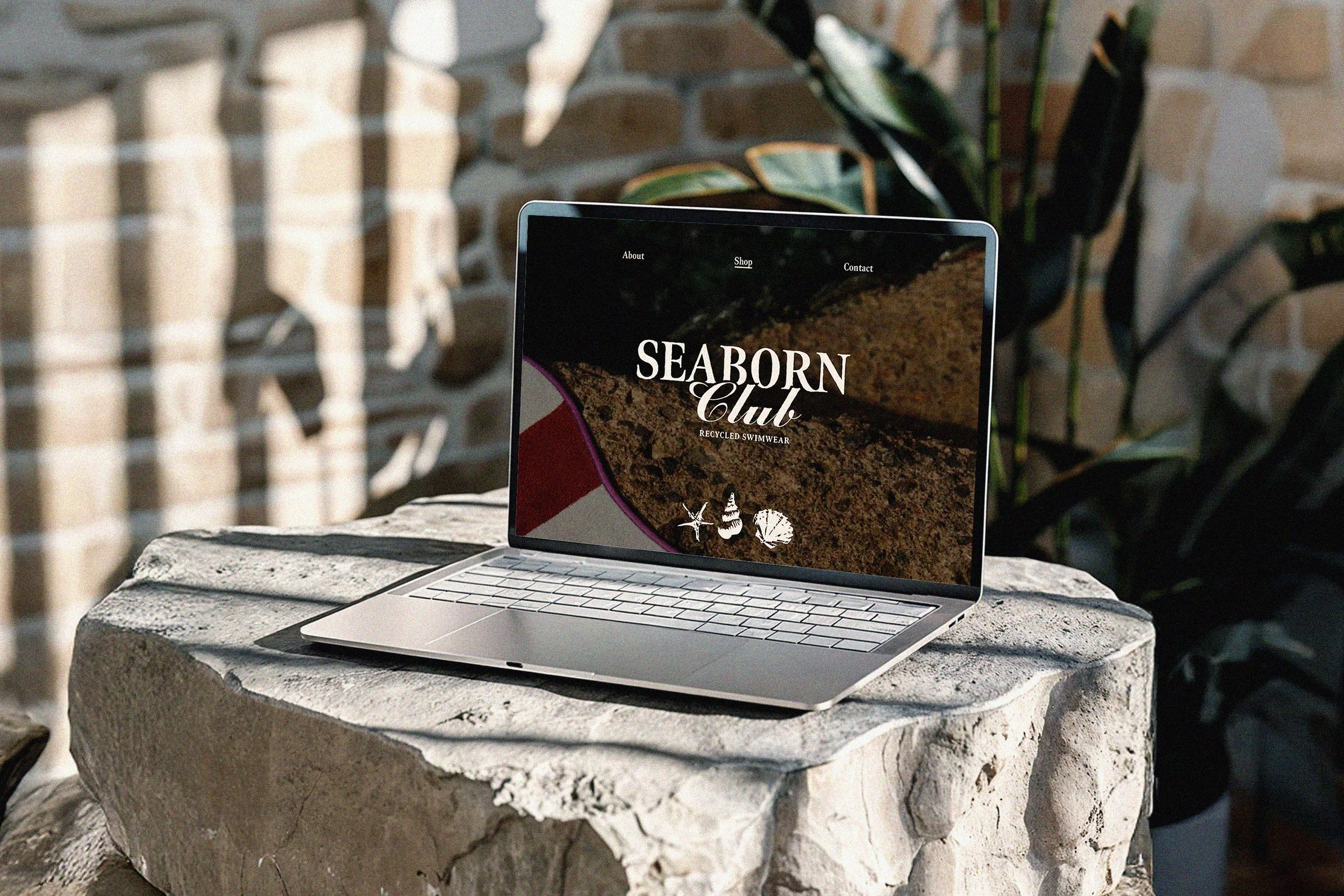 A laptop on a stone table outdoors displaying the website for Seaborn Club, a recycled swimwear brand, with a background of a brick wall and green plants.