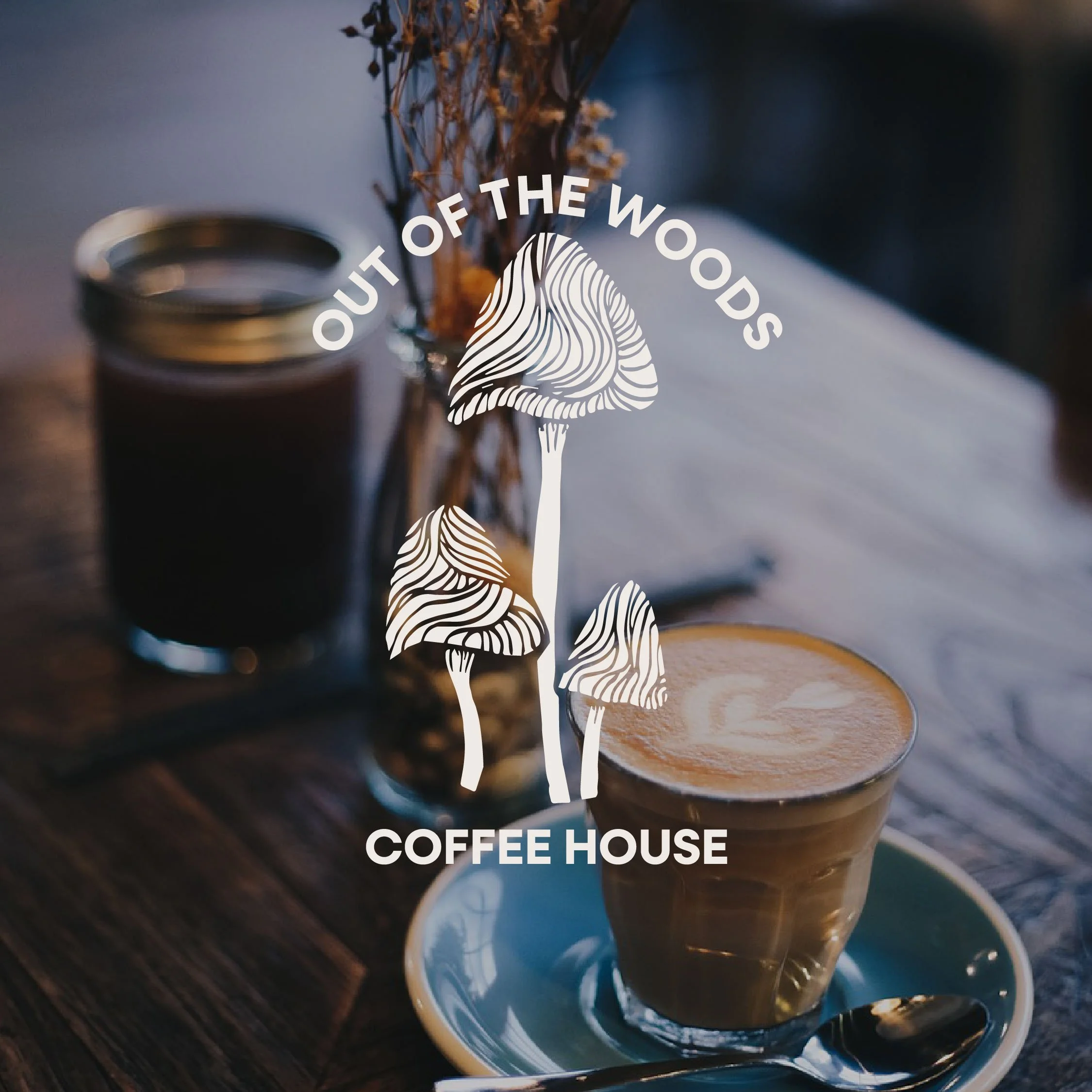A close-up of a cup of coffee on a saucer with a spoon, with a jar of coffee and a vase with flowers in the background, overlaid with a logo that says 'Out of the Woods Coffee House' with mushrooms illustration.
