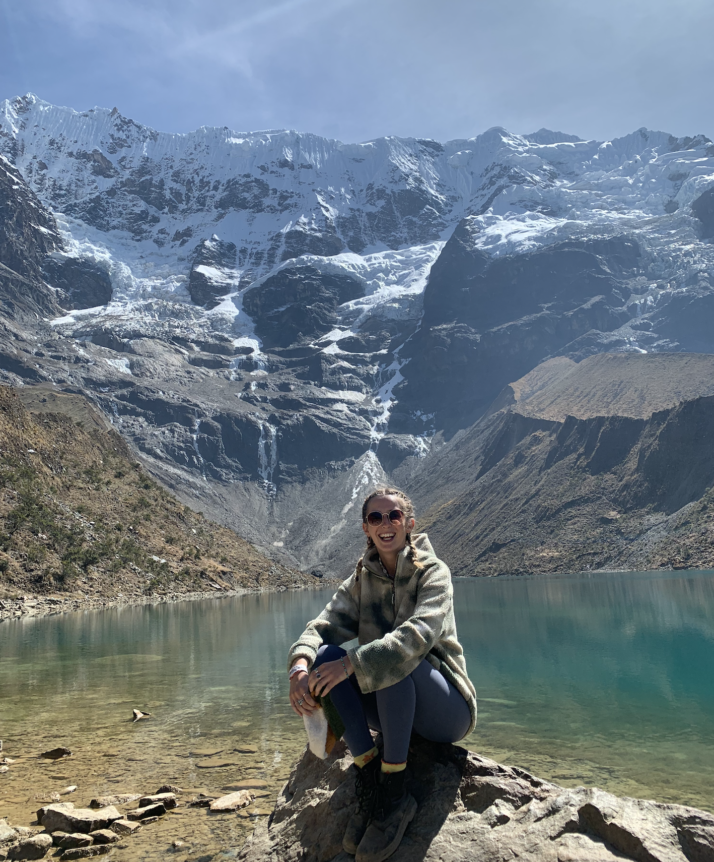 A woman sitting on a rock near a clear lake, with snow-capped mountains in the background. She is wearing sunglasses, a fleece jacket, and has braided hair, smiling at the camera.