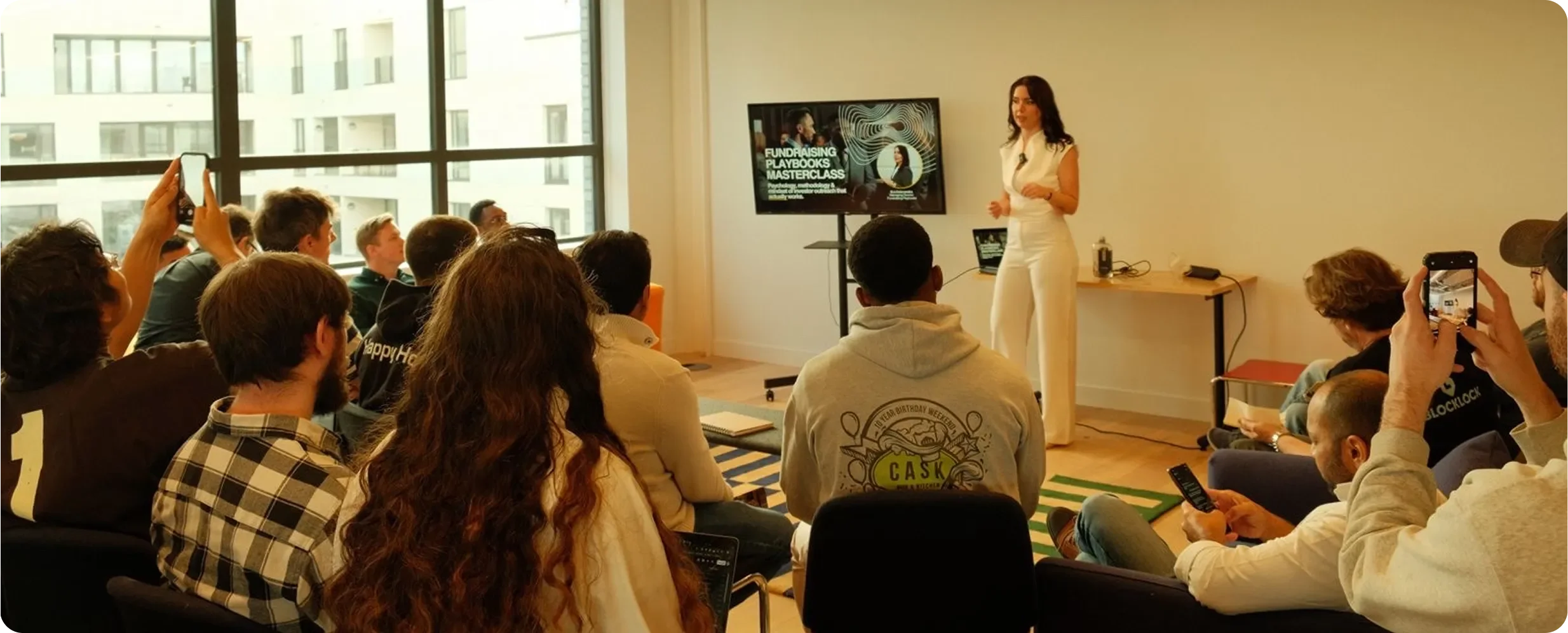 A woman giving a presentation to an audience in a room with large windows. The presentation slide reads 'Fundraising Playbooks Masterclass.' Several audience members are taking photos or videos with their smartphones.