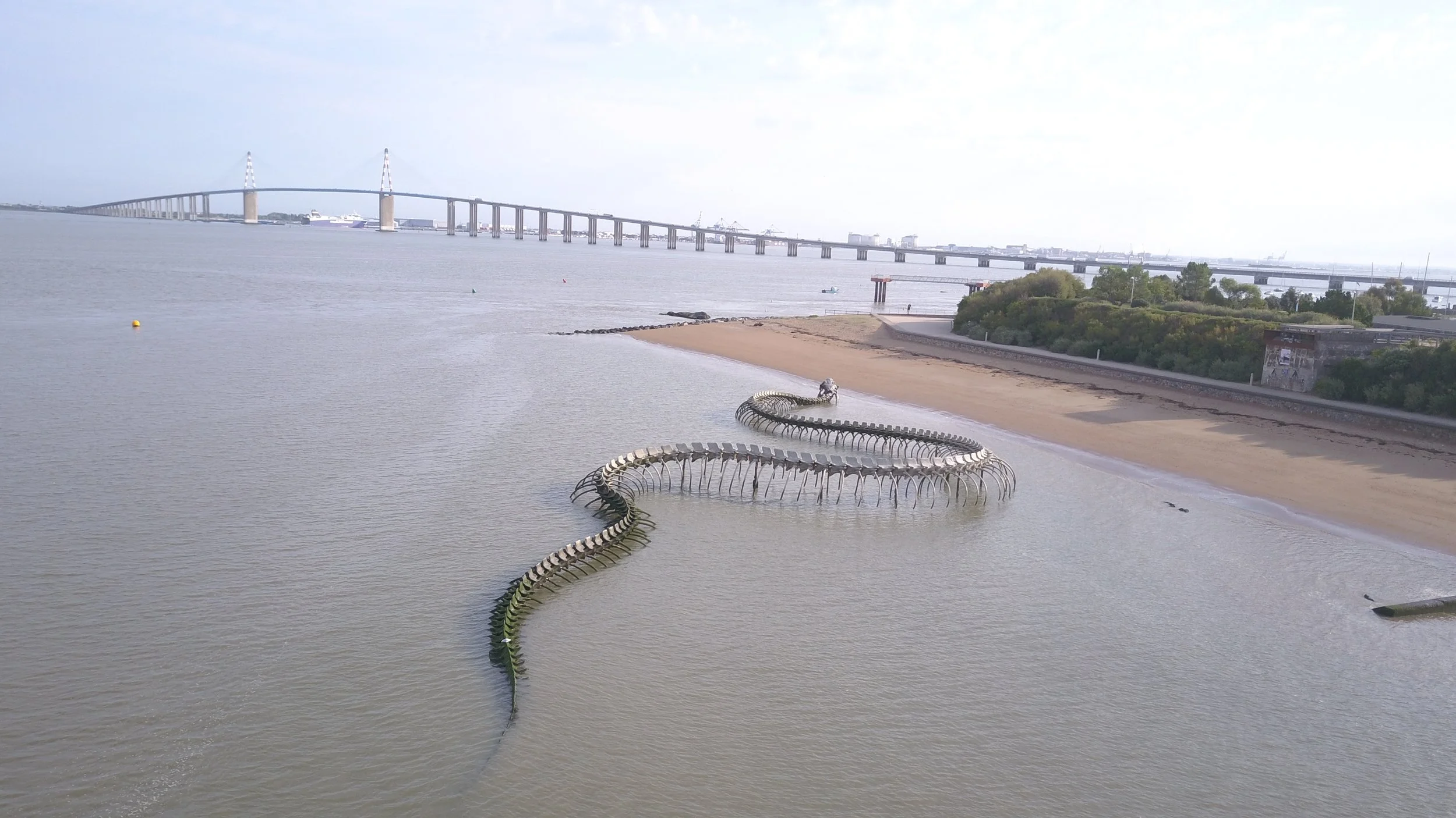 Sculpture d'un serpent en métal situé sur une plage au bord de l'eau avec un pont en arrière-plan.