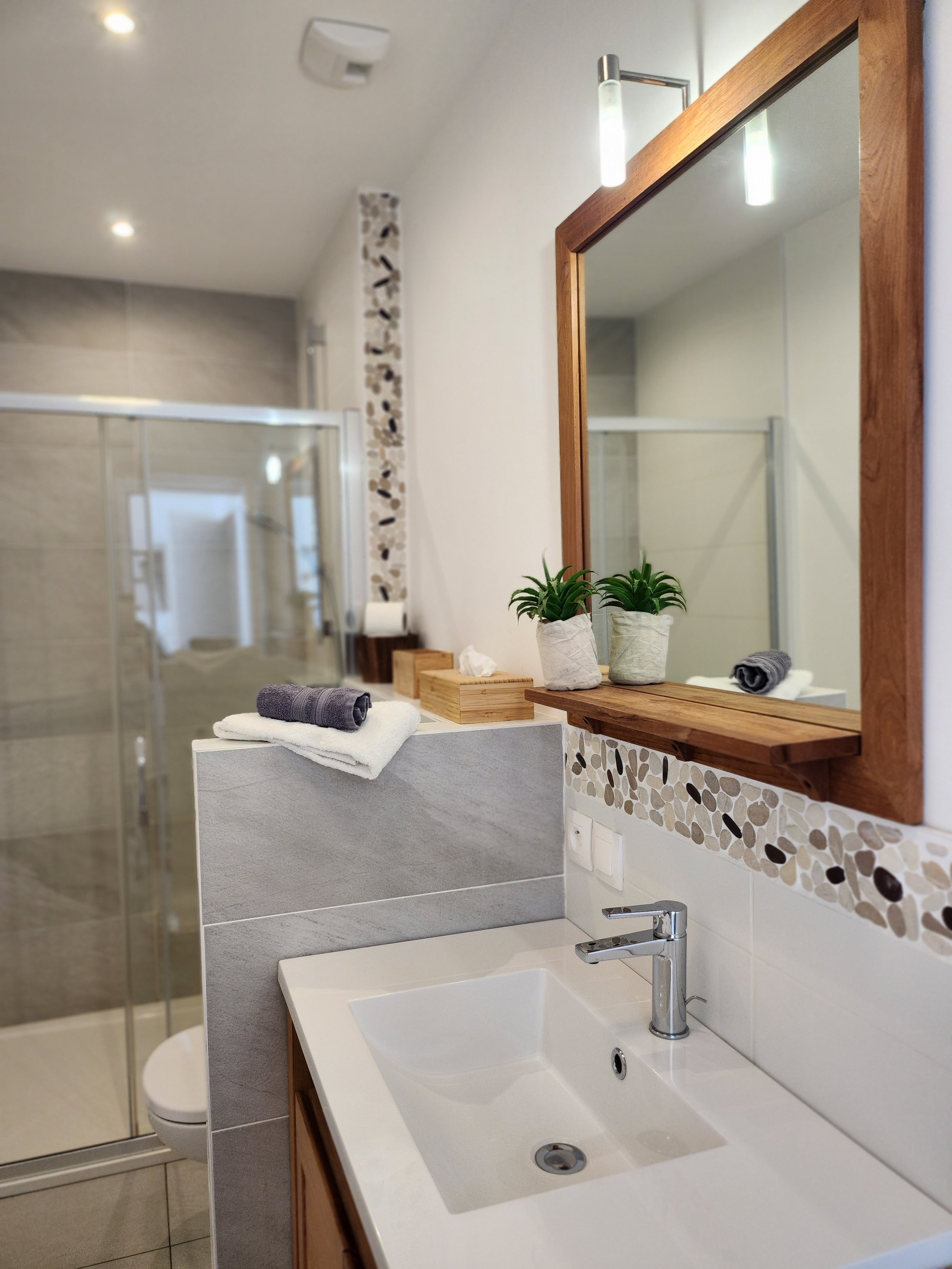 Salle de bains moderne avec lavabo, miroir en bois, plantes, serviettes et coin douche.