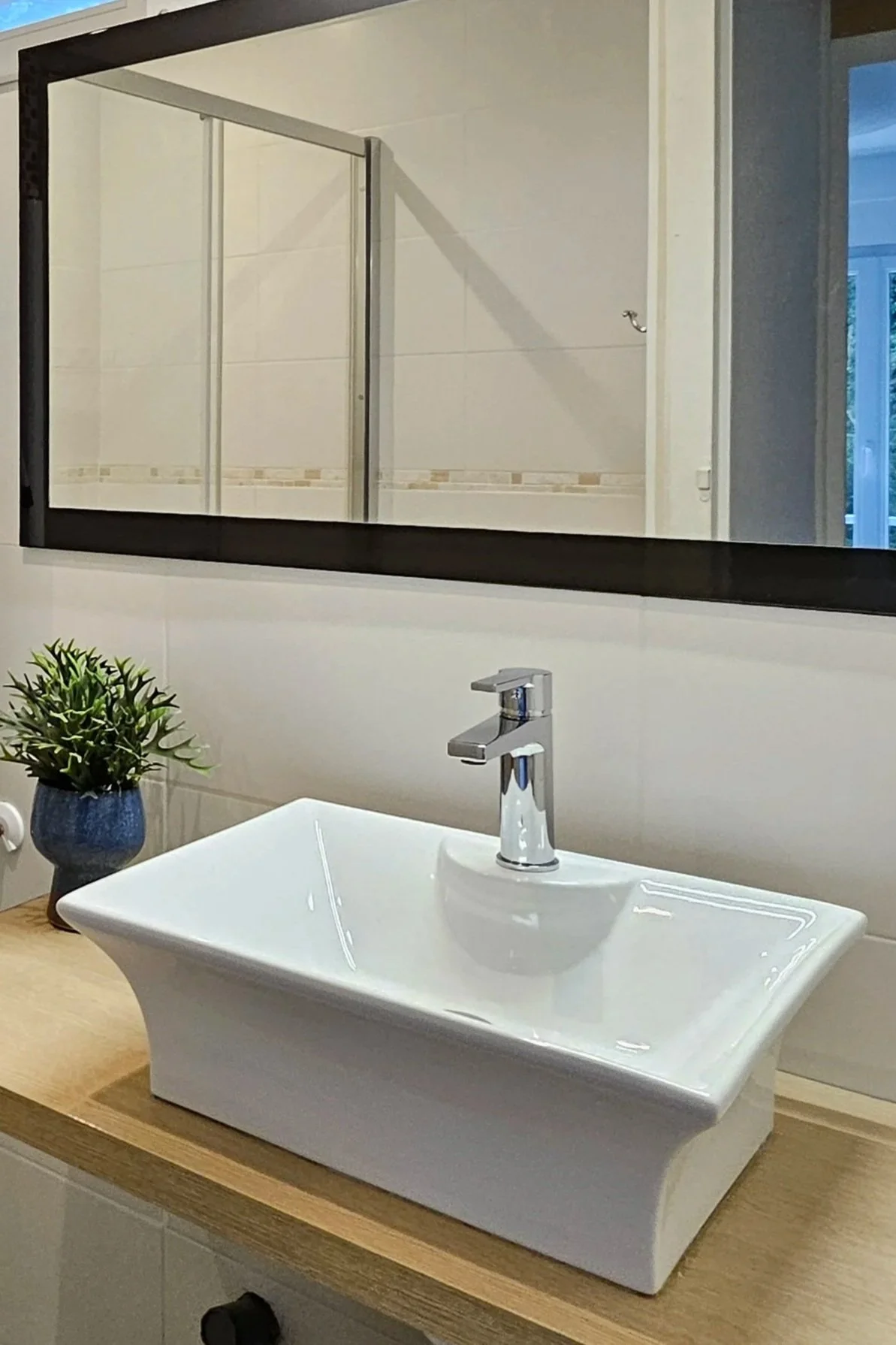 Un lavabo blanc moderne avec robinet en métal, placé sur une surface en bois dans une salle de bain. À gauche, une plante verte dans un pot bleu, un miroir rectangulaire au-dessus du lavabo. En arrière-plan, une douche avec parois en verre et une porte ouverte laissant entrer de la lumière naturelle.