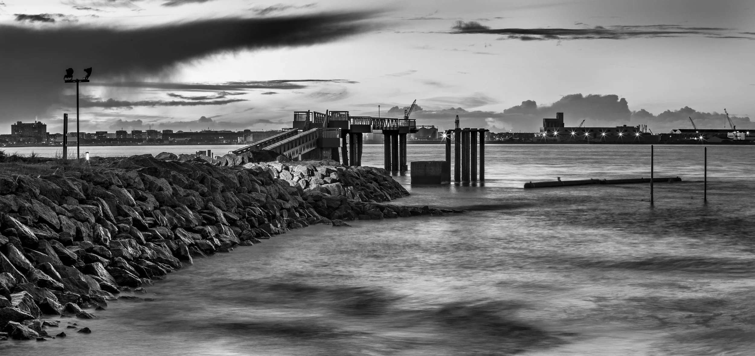 Vue en noir et blanc de la jetée quartier Mindin, sur l'estuaire de la Loire, en premier plan amas de rochers avec une plateforme, et ville de Saint-Nazaire  et chantiers atlantiques en arrière-plan, sous un ciel nuageux.