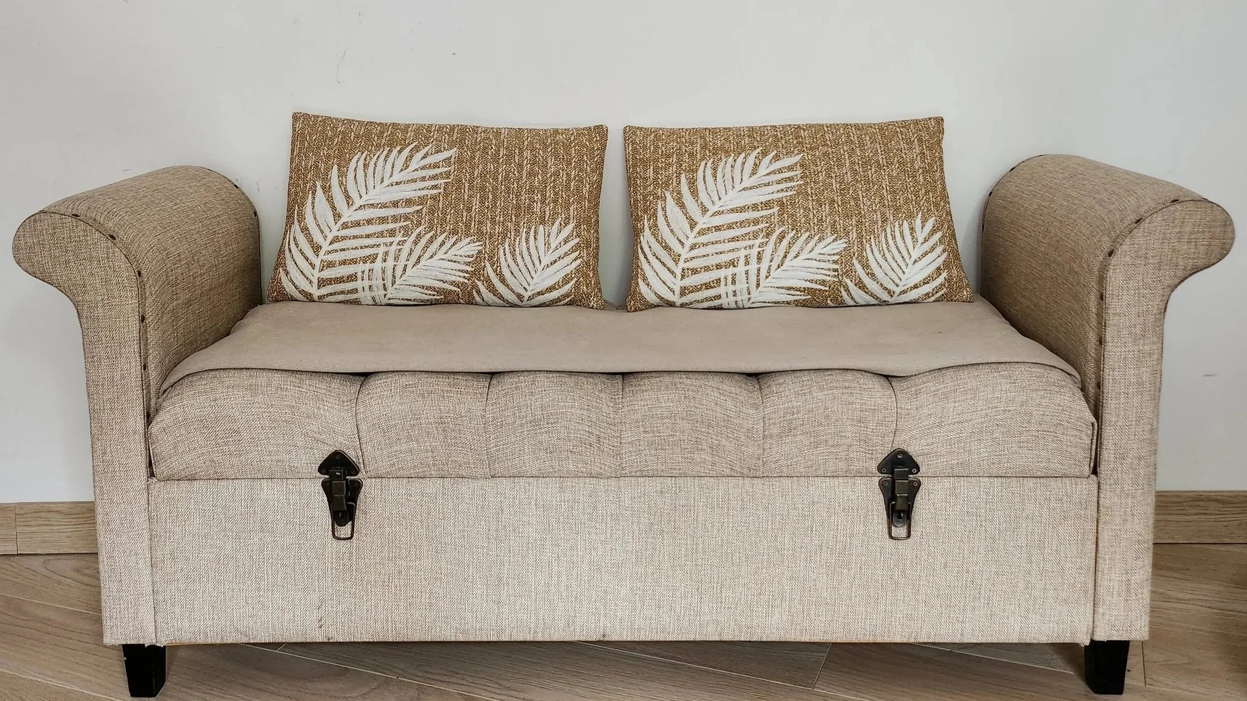 Canapé beige avec deux coussins à motif de feuilles blanches, boutons de fixation visibles à l'avant, posé contre un mur blanc.