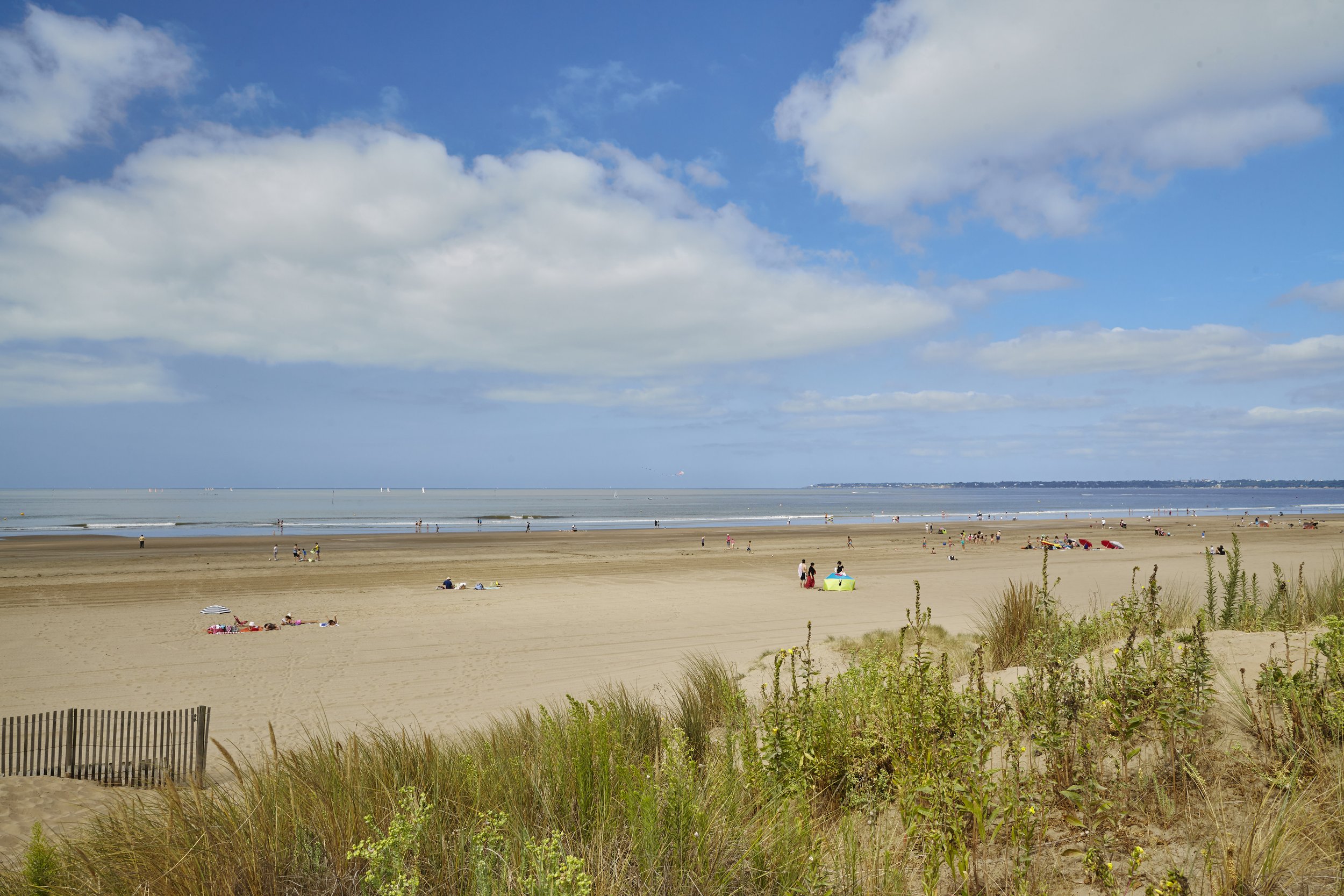 Plage de sable avec des personnes se baignant, allongées, et jouant, sous un ciel partiellement nuageux, avec des herbes et une clôture en bois au premier plan.