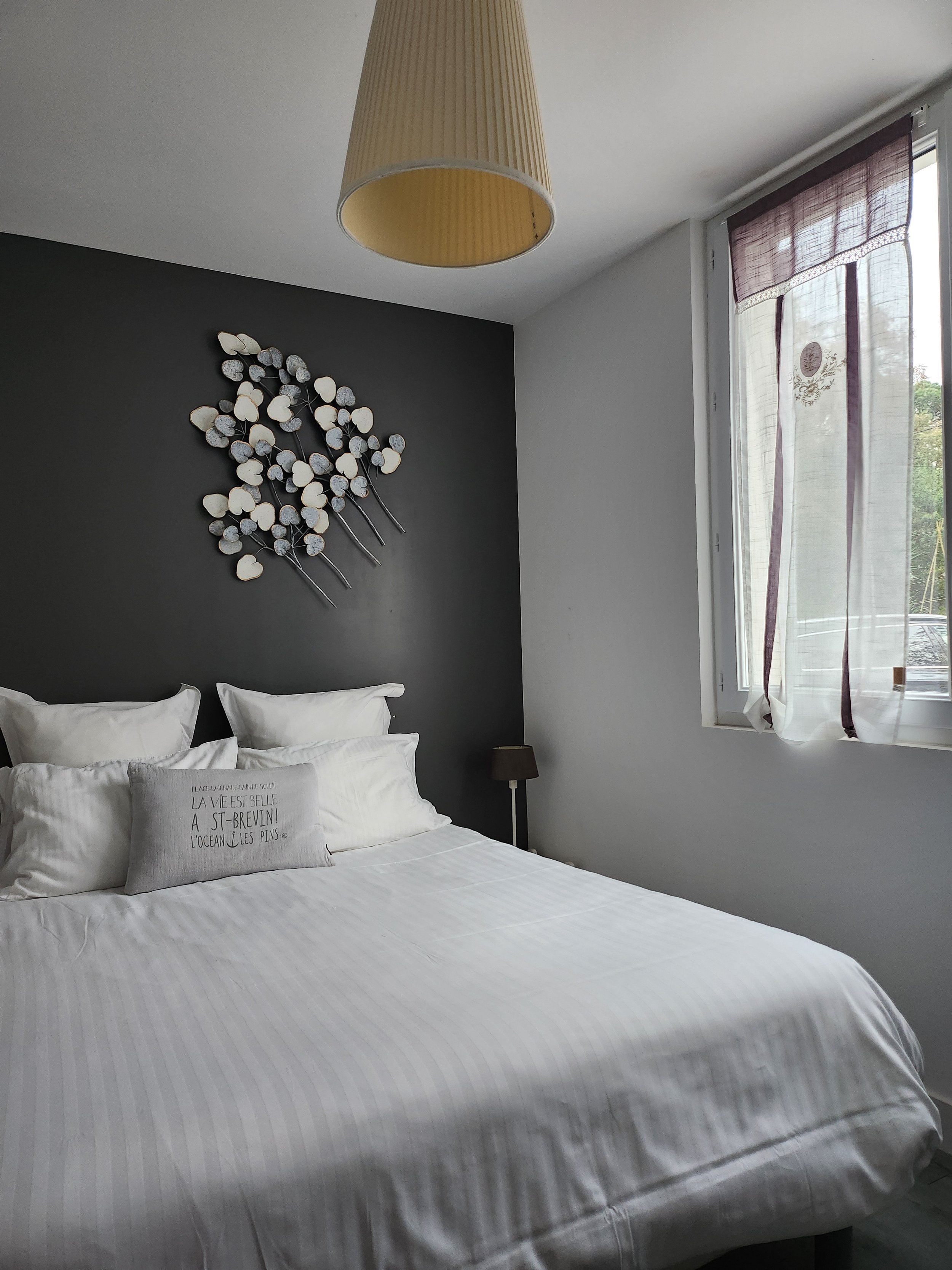 Chambre à coucher avec un lit blanc, des oreillers et un coussin avec une inscription. Mur noir au-dessus du lit avec une décoration murale d'artefacts en forme de fleurs. Fenêtre avec rideau léger violet et vue extérieure.