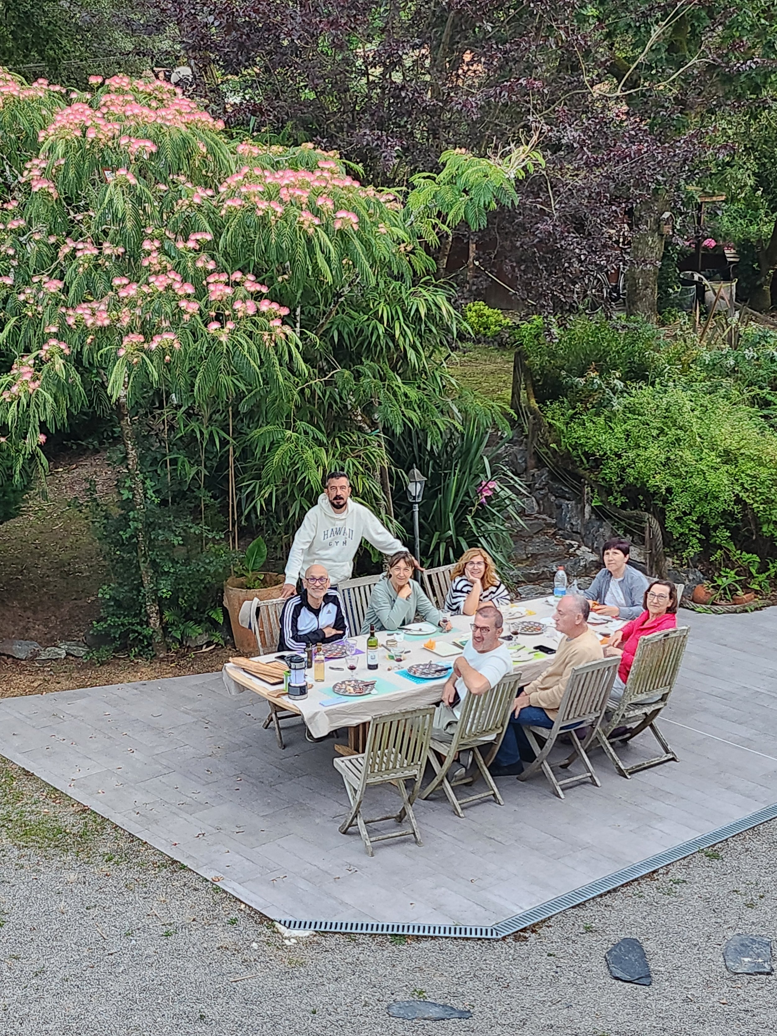 Groupe de huit personnes assises autour d'une table d'extérieur pour un repas, avec un homme debout derrière eux. La scène se déroule dans un jardin avec de grandes plantes vertes et des arbres en arrière-plan.