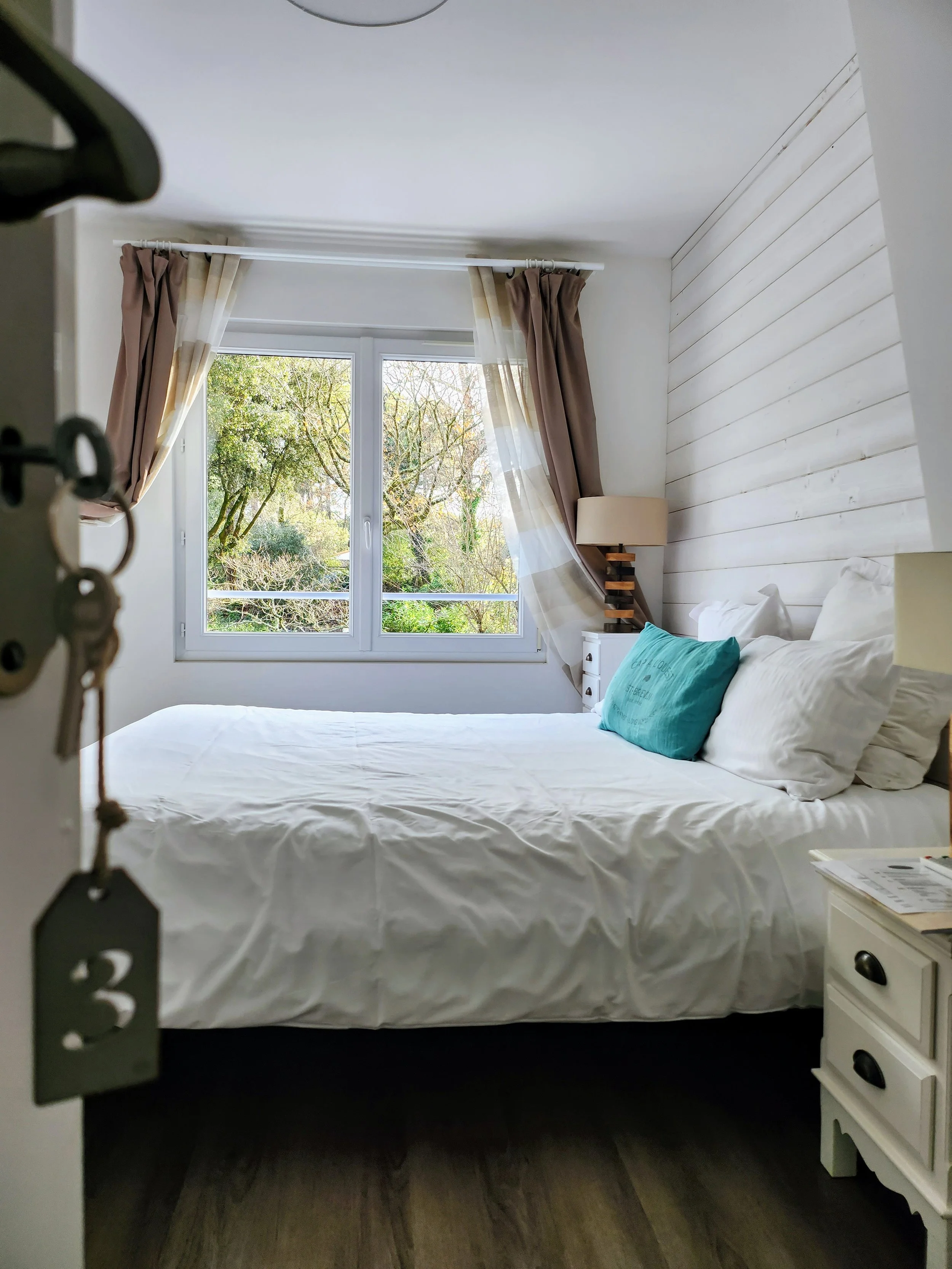 Chambre avec lit enroulé, vue sur une fenêtre avec des arbres dehors, draps blancs, coussins, lampe de chevet et décoration boisée.