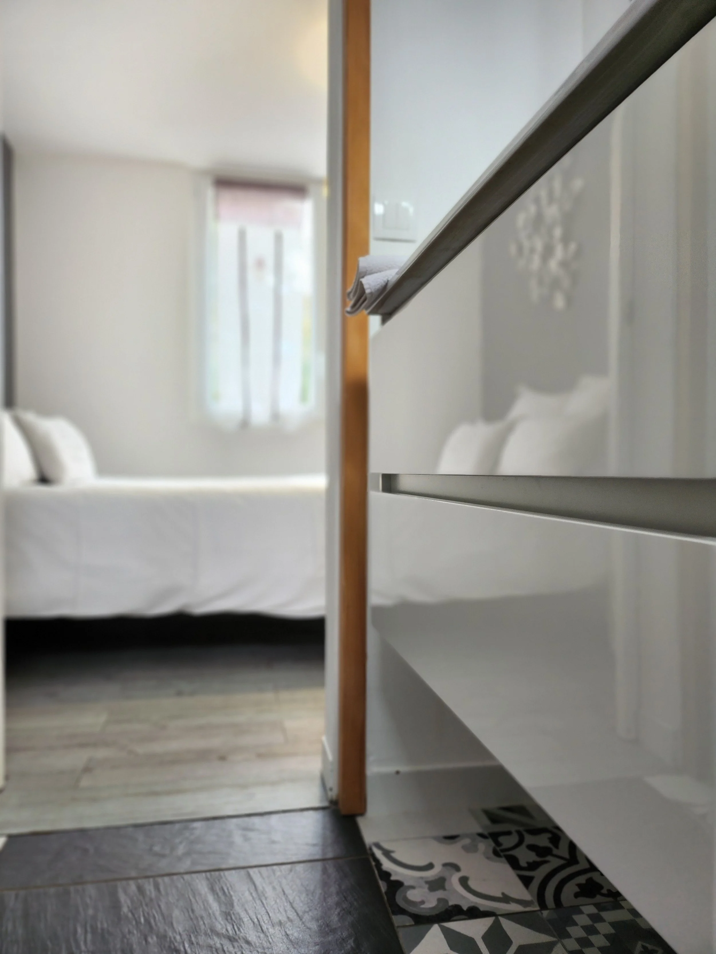 Vue partielle d'une chambre avec un lit blanc, un mur avec une fenêtre à rideaux, un parquet en bois clair, une commode blanche, et un sol en carrelage avec un motif noir et blanc.