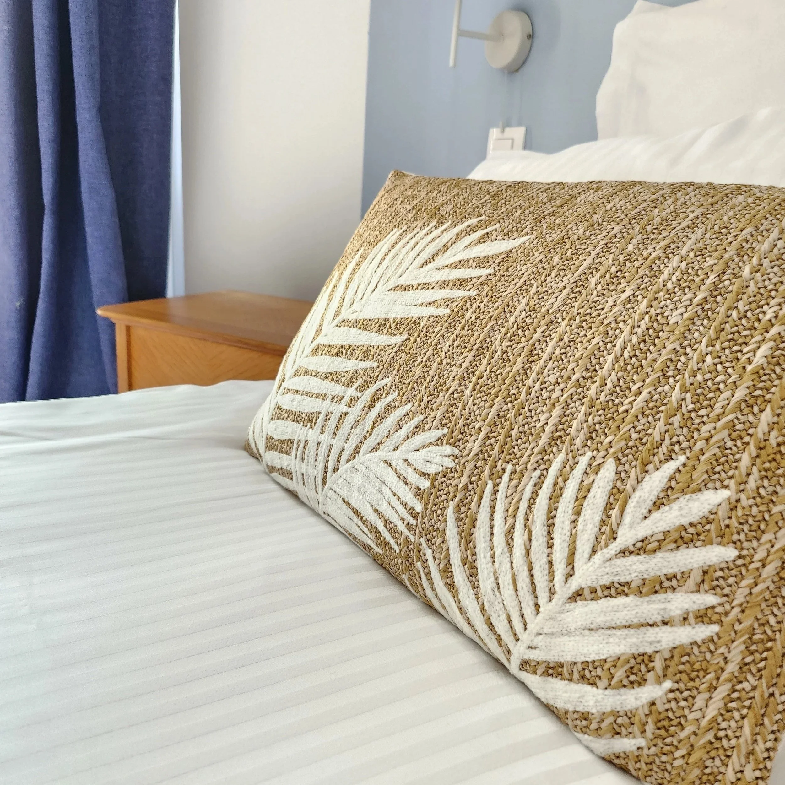 Coussin beige avec motif de feuilles blanches sur un lit avec drap blanc, à côté d'une table en bois, dans une chambre avec rideaux bleus.