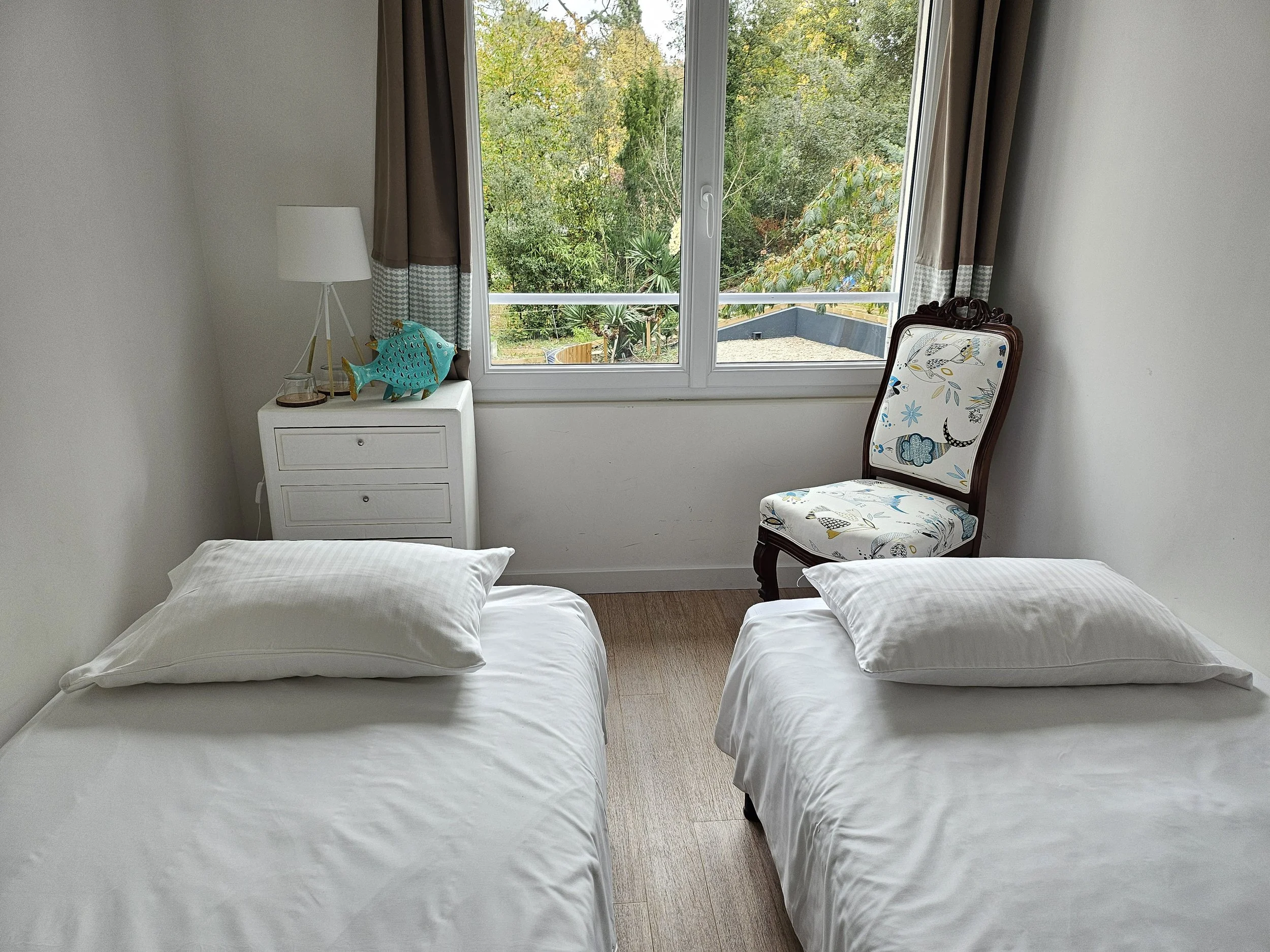 Chambre d'hôte annexe de la suite familiale brevocean avec deux lits simples, une fenêtre donnant sur jardin, une petite commode, une lampe, une décoration poisson, un fauteuil avec un tissu à motif floral.