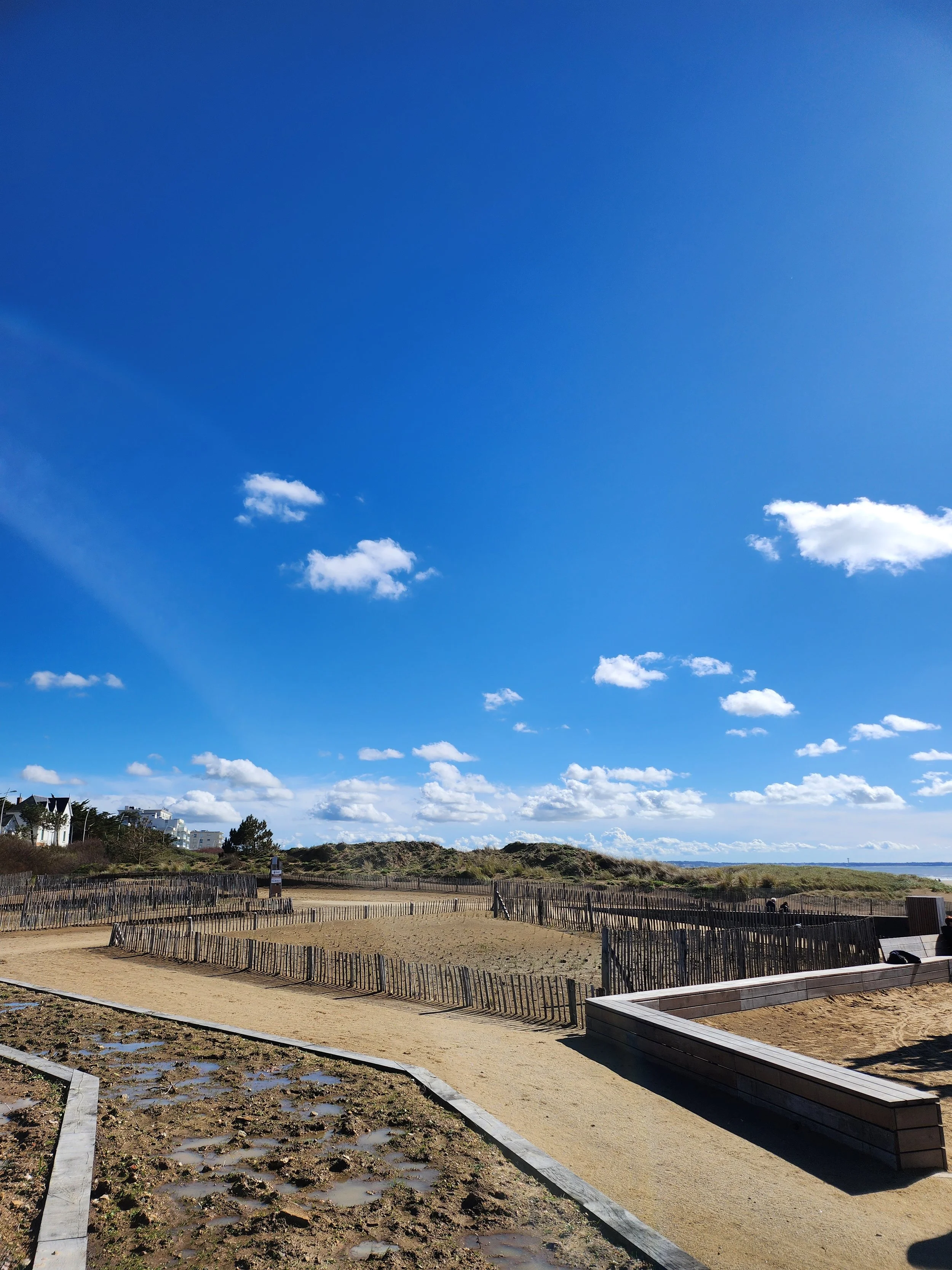Plage avec sable, dunes, ciel bleu avec quelques nuages, chemin de sable et clôtures en bois et jeux d'enfants sur le littoral atlantique.