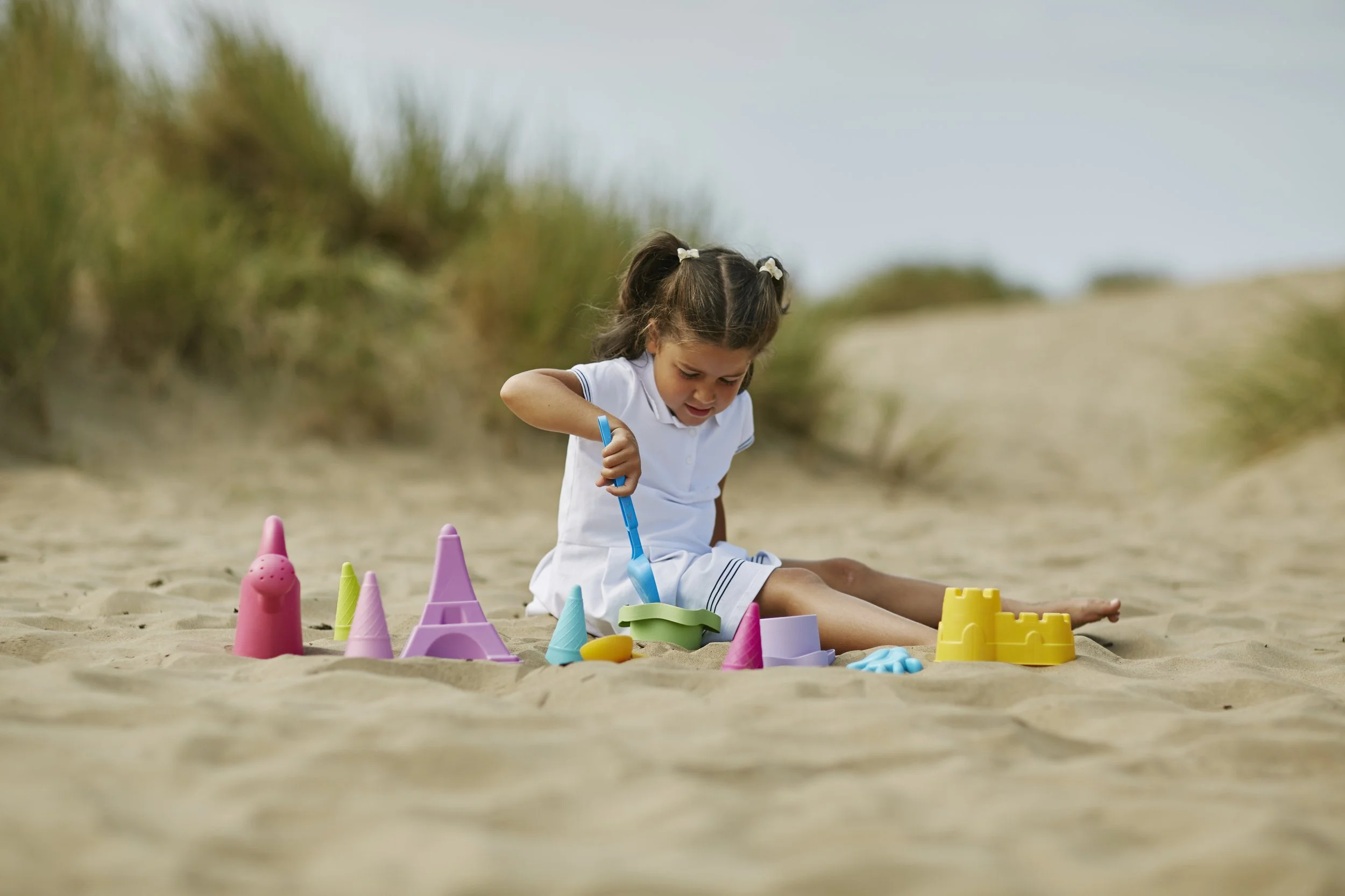 Une jeune fille joue seule sur la plage avec des jouets de sable colorés, notamment des châteaux, des pelles et des seaux, avec des dunes et de la végétation en arrière-plan.
