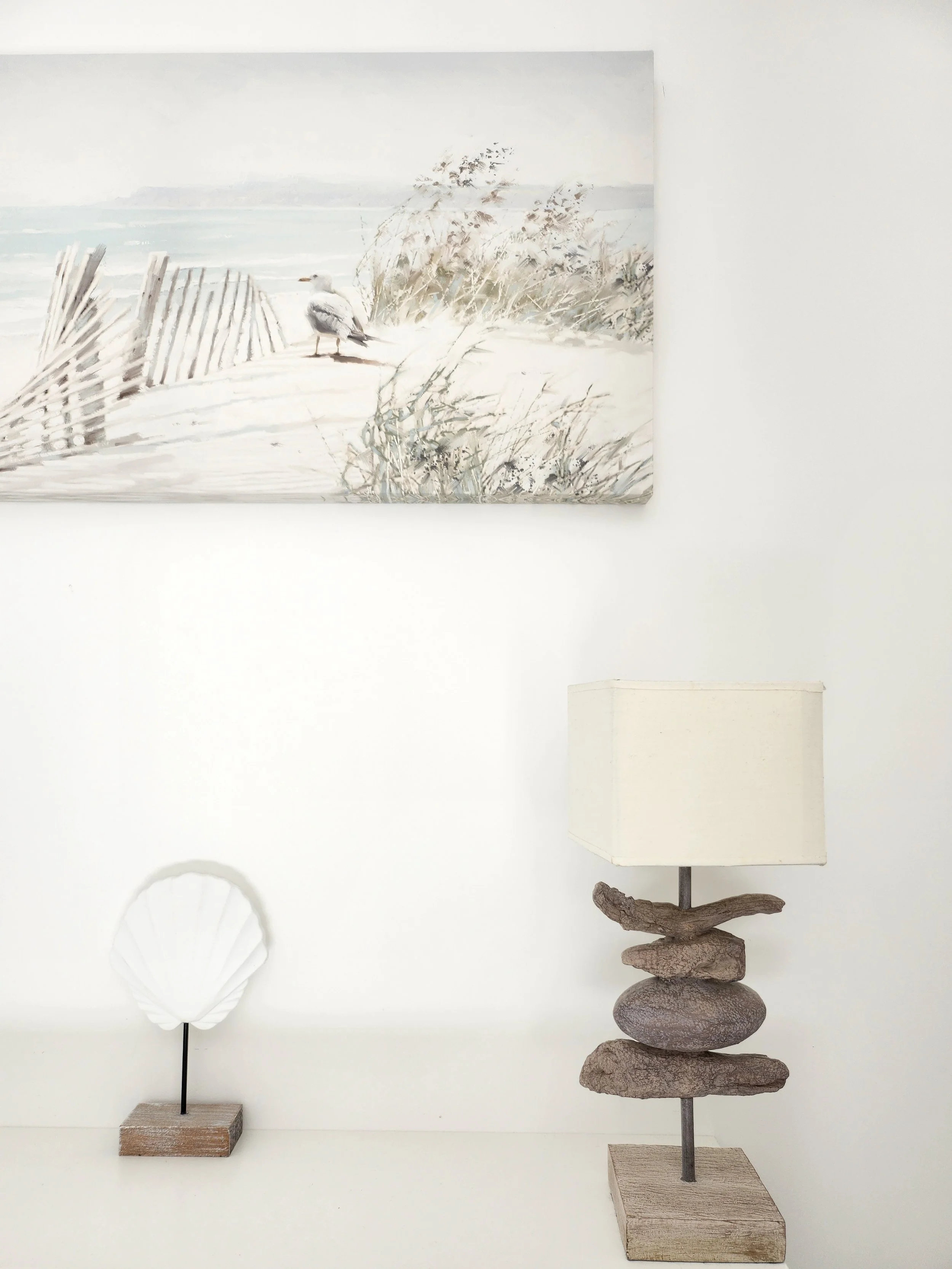 Une lampe de table avec un pied en pierres naturelles empilées et un abat-jour blanc, et une petite sculpture en forme de coquillage blanc sur une base en bois, placé devant un mur blanc avec une peinture représentant une scène de plage avec un oiseau, des dunes de sable et de la végétation
