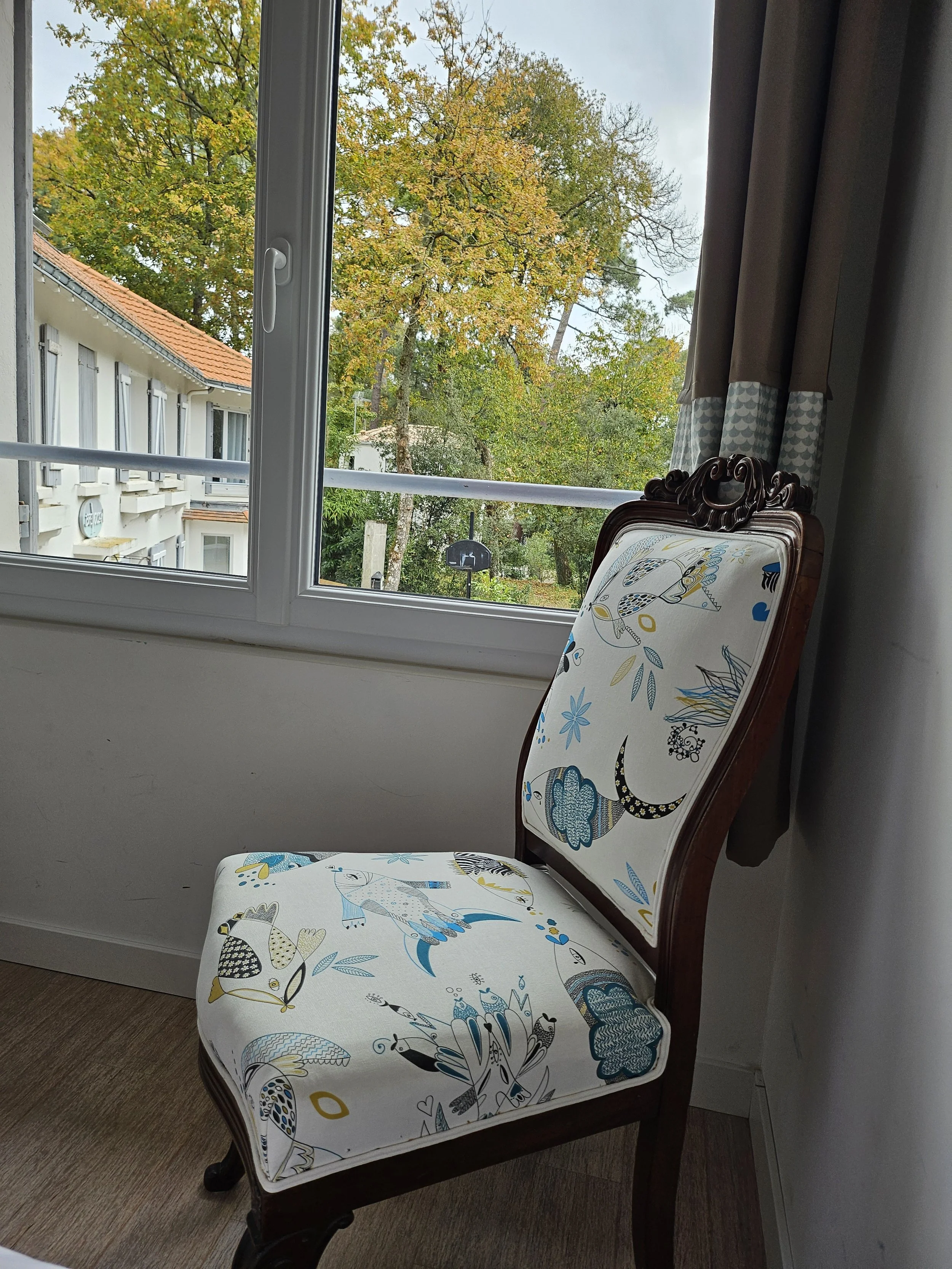 Chaise ancienne en bois avec un tissu à motifs colorés blanc, bleu, gris et or, près d'une fenêtre laissant apparaître des arbres avec des feuilles vertes et jaune, et un bâtiment avec un toit en tuiles rouges.