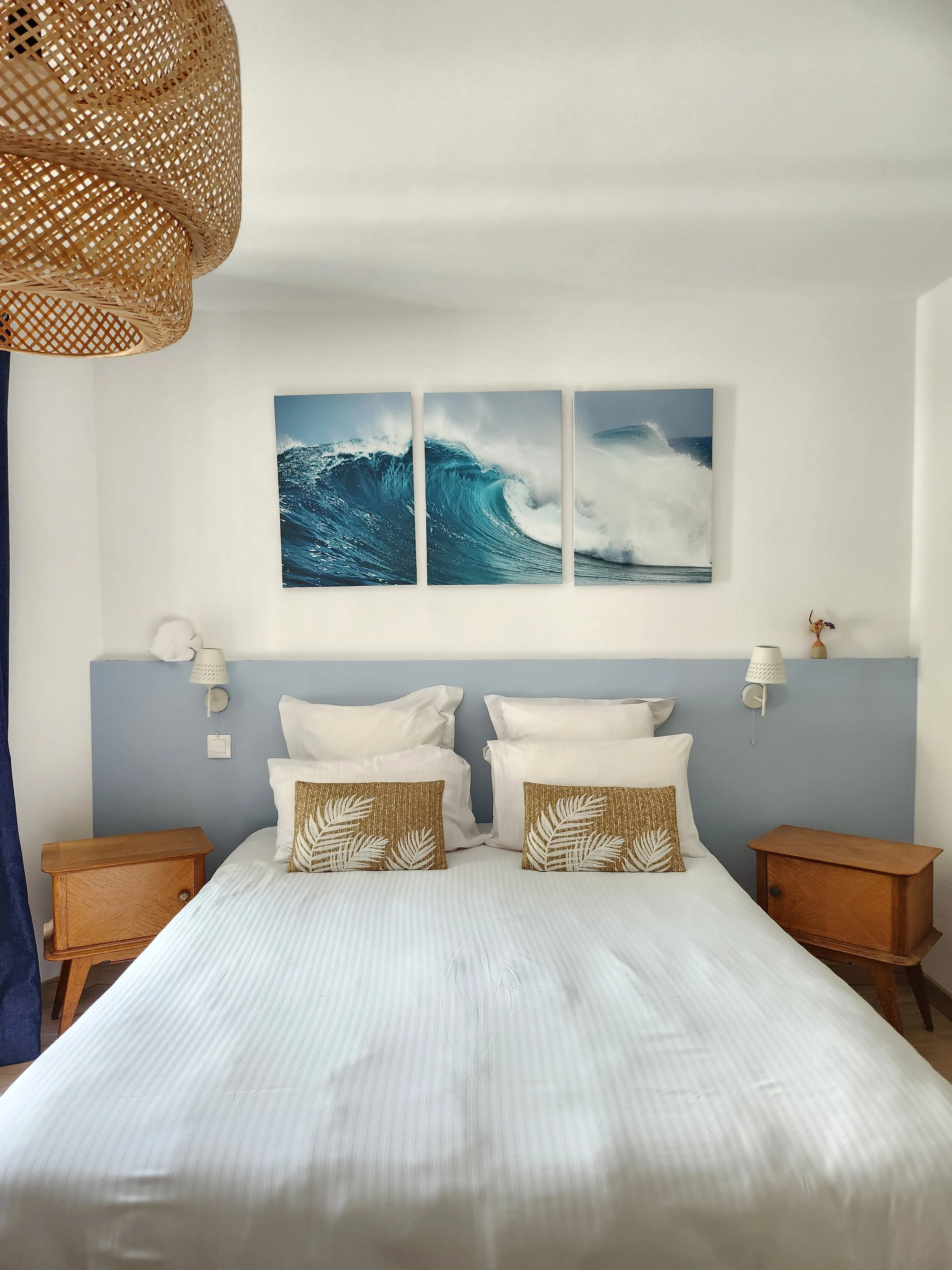 Chambre moderne avec tête de lit bleue, tableau triptyque représentant des vagues, deux lampes murales, deux tables de nuit en bois, et oreillers avec housses à motifs de feuilles.