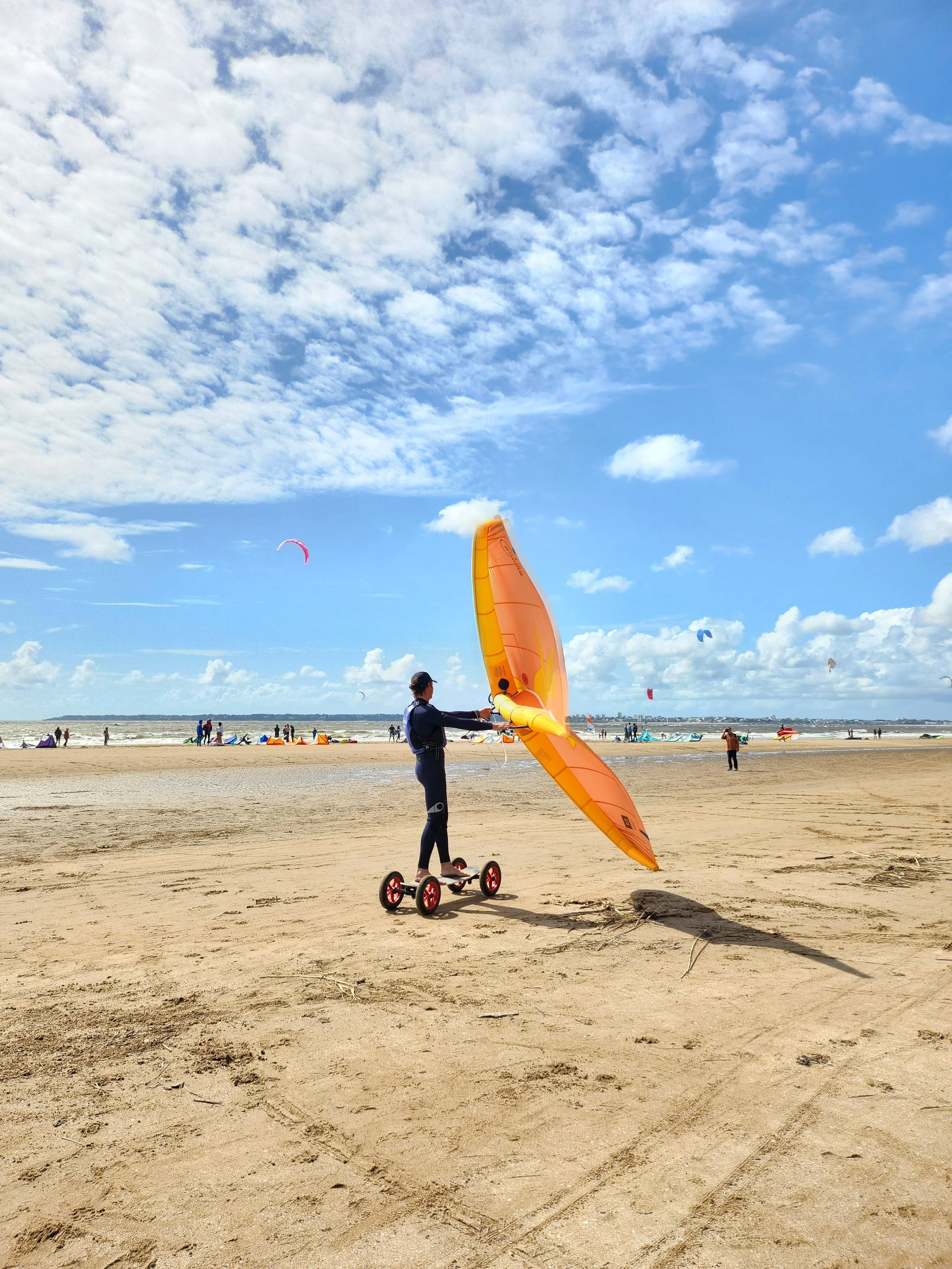 Homme pratiquant un sport de glisse sur une plage en bord de mer à Saint-Brevin-les-Pins, sur un mountainboard tenant une voile de wingfoil orange  avec un ciel bleu et des nuages, cerfs-volants dans le ciel et des gens au loin.