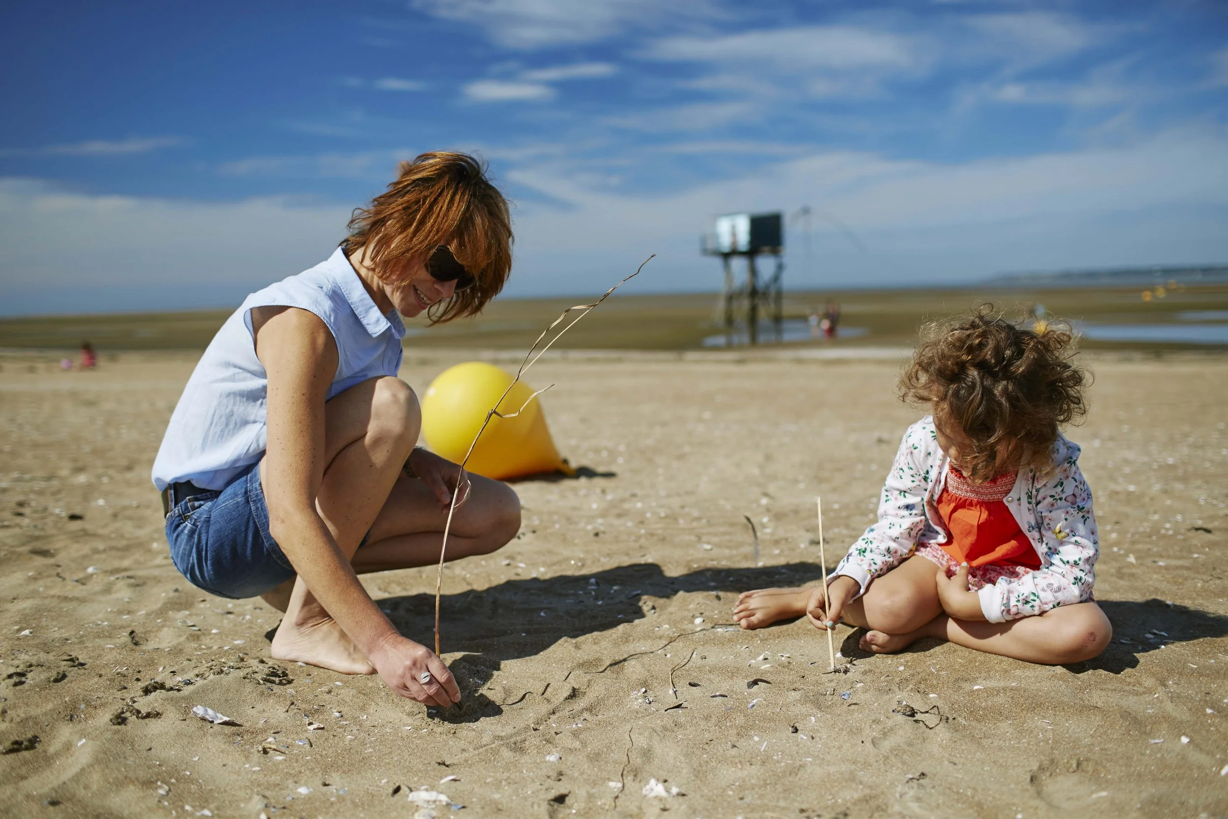 Une femme et une jeune fille en vacances en loire-atlantique jouent à la pêche ou au chasse au trésor sur la plage, assises dans le sable avec des bâtons et des fils. Le ciel est bleu avec quelques nuages, et il y a un grand flotteur jaune derrière e