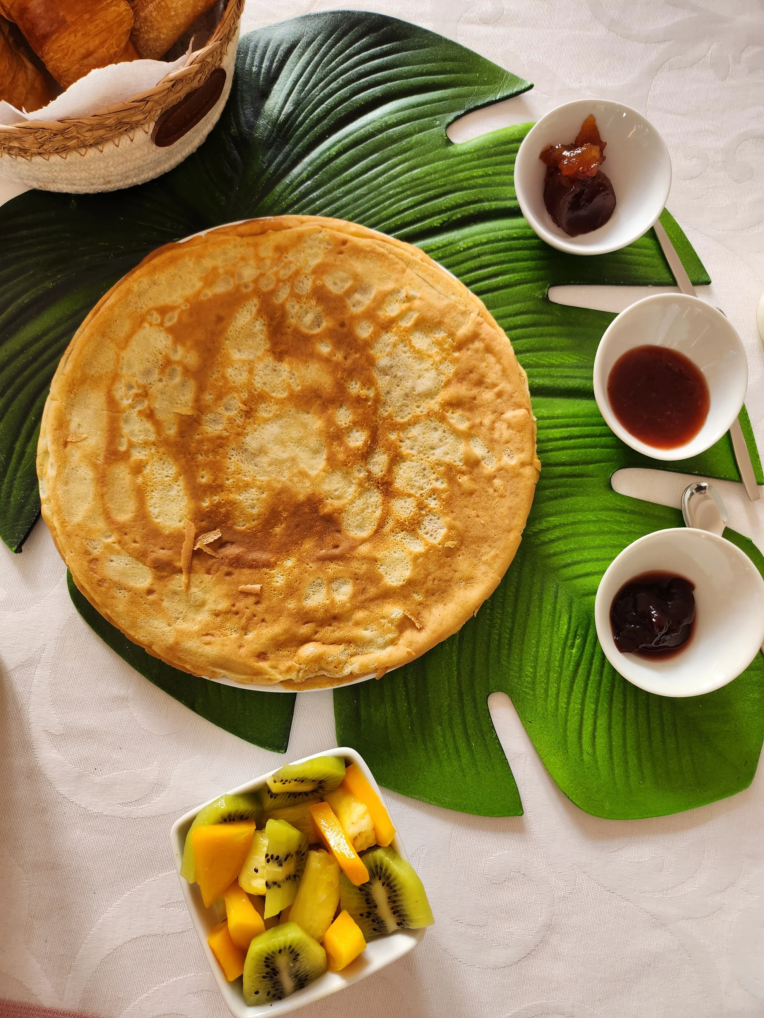 Crêpe servie au petit-déjeuner de la chambre d'hôtel, avec un bol de fruits coupés comprenant du kiwi et de la mangue, et trois petits ramequins contenant des confitures maison, sur une nappe blanche.