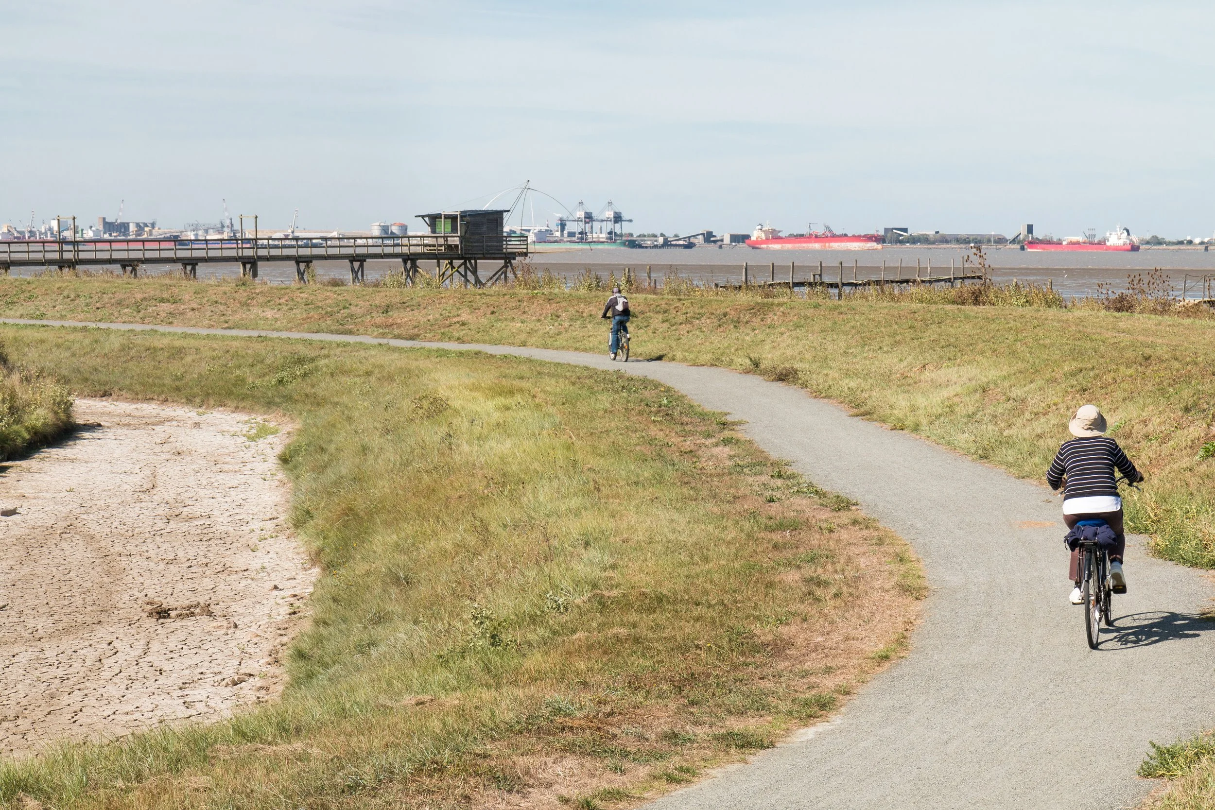 Deux personnes font du vélo sur un chemin de terre et un sentier en bord de mer avec un port et des bateaux en arrière-plan.